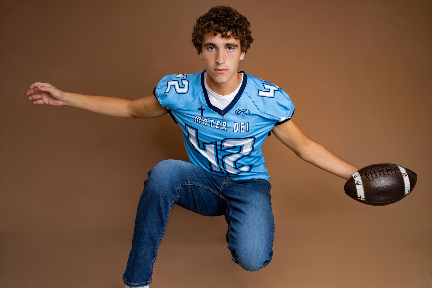 High school football player in light blue jersey and jeans poses with football against brown backdrop in studio setting.
