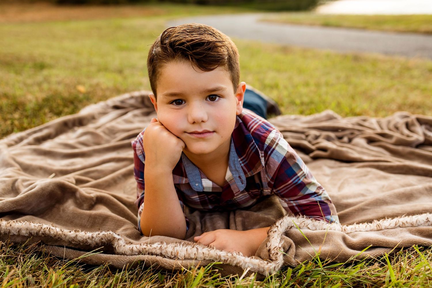 Child laying on a brown blanket outdoors on grass wearing plaid shirt smiles at camera during sunny day.