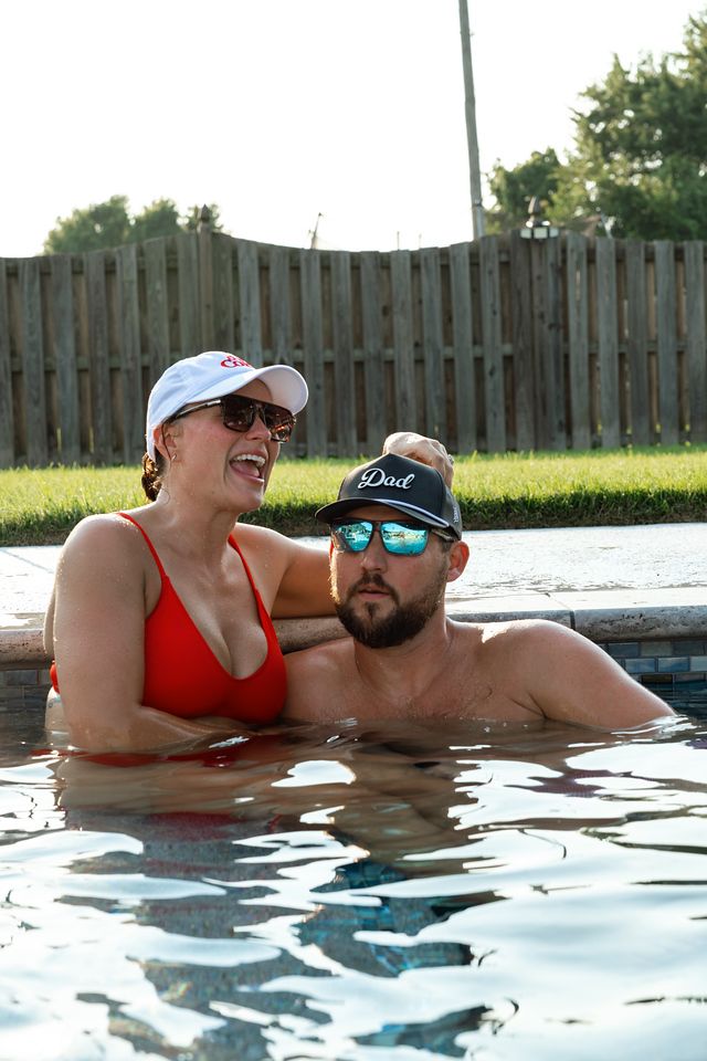 Couple relaxing together in an outdoor pool, woman in red bikini and white cap, man wearing a Dad hat and sunglasses.