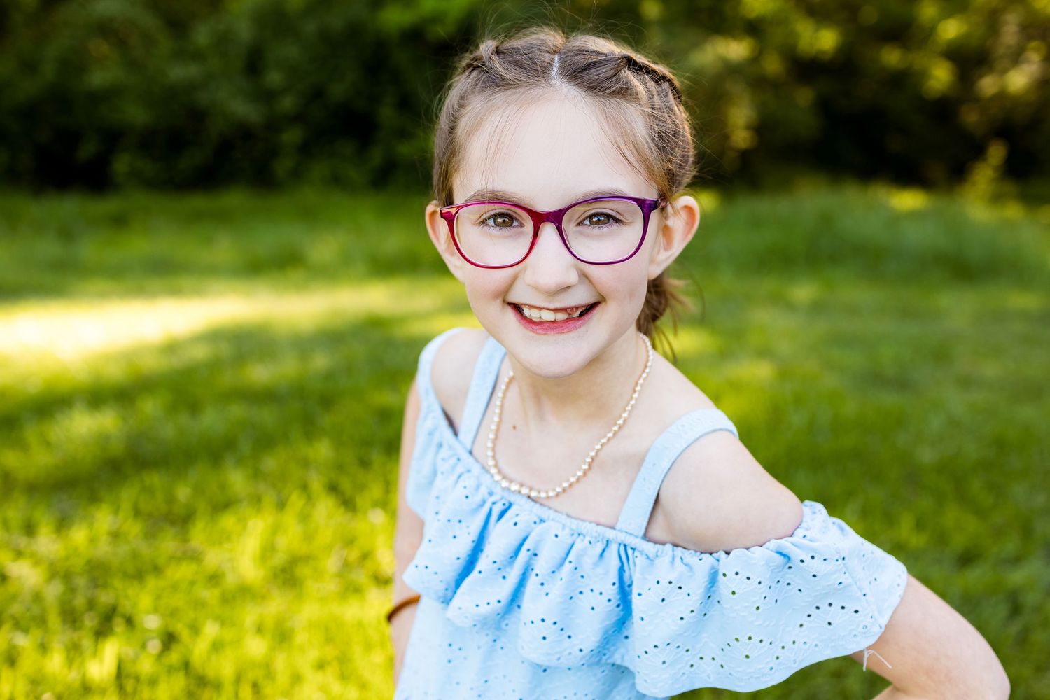 Happy child in blue eyeglasses and ruffled dress poses playfully in sunny garden setting.
