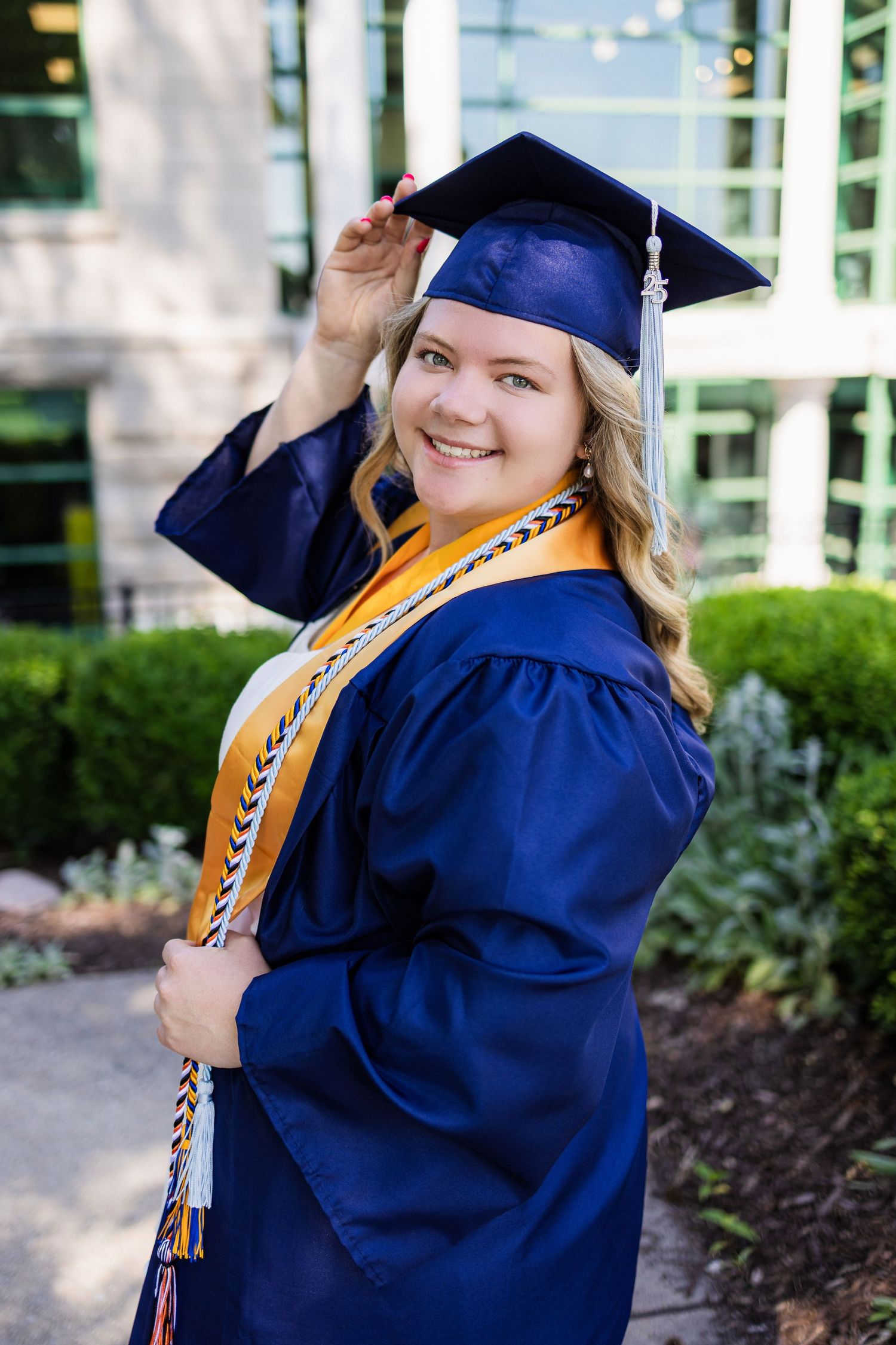 A graduate in blue cap and gown with yellow honors cords poses outdoors on campus.