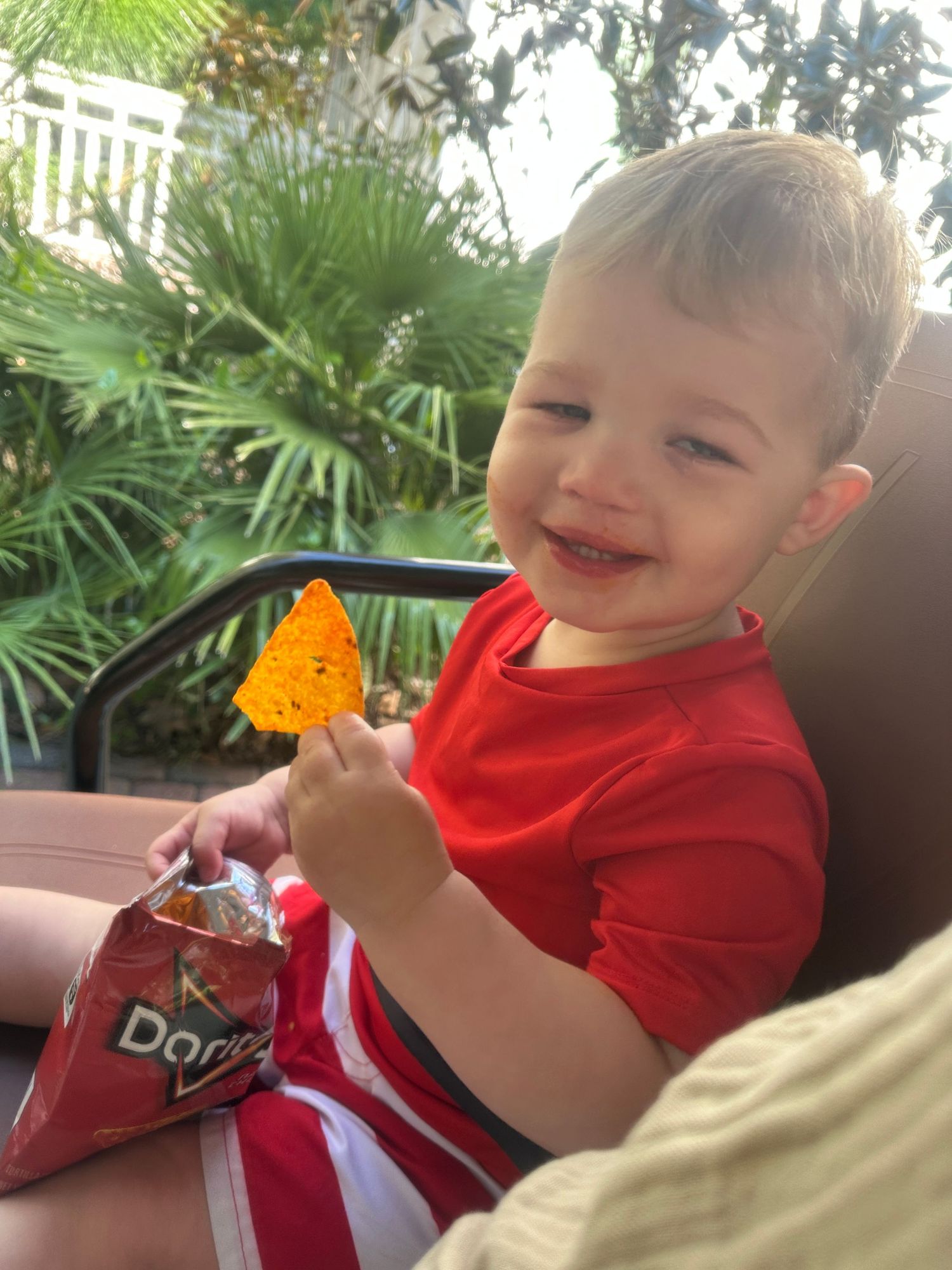 Child smiling while holding orange snack with tropical plants in background.