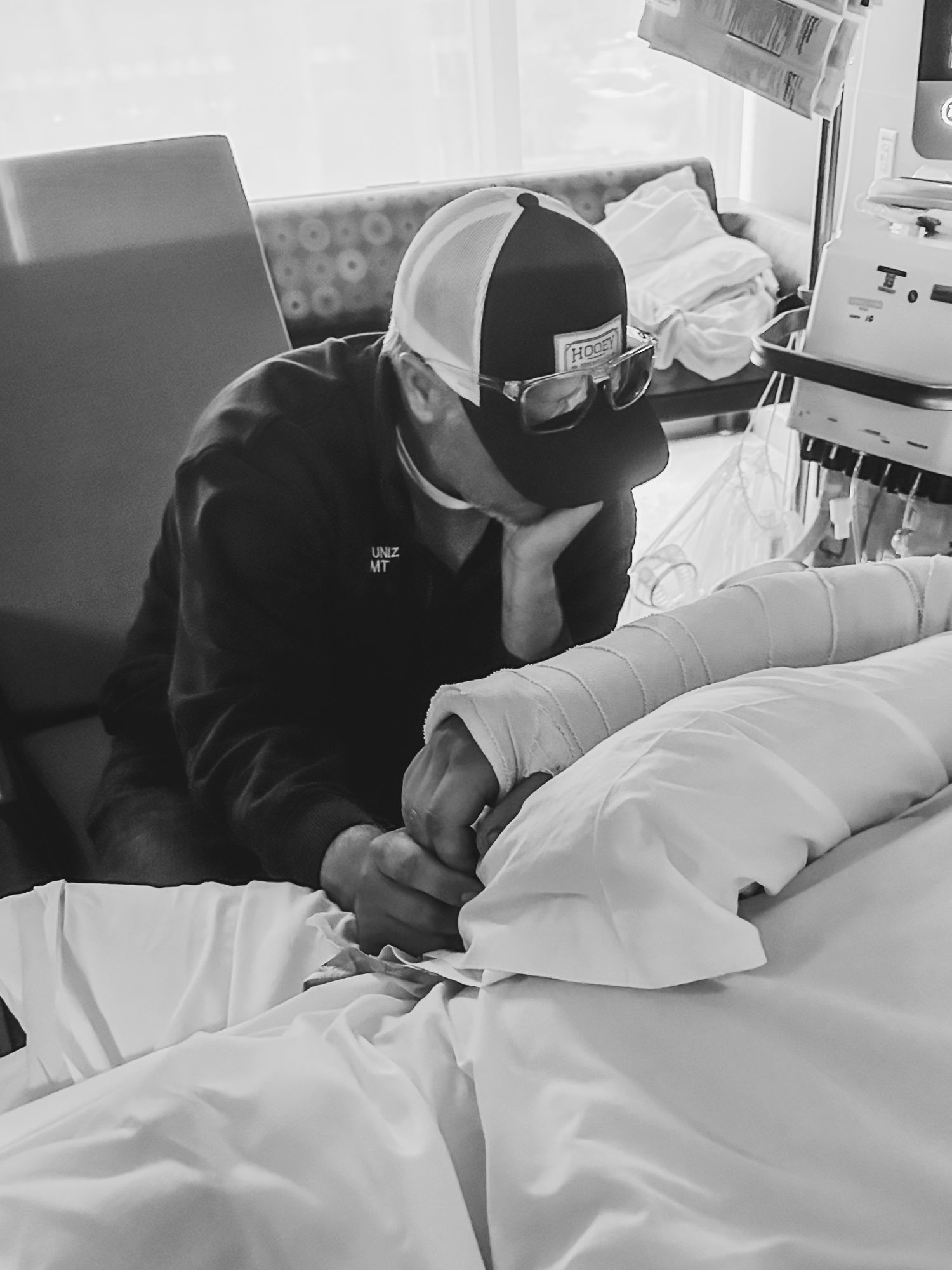Person in baseball cap sits beside hospital bed holding hand of patient.
