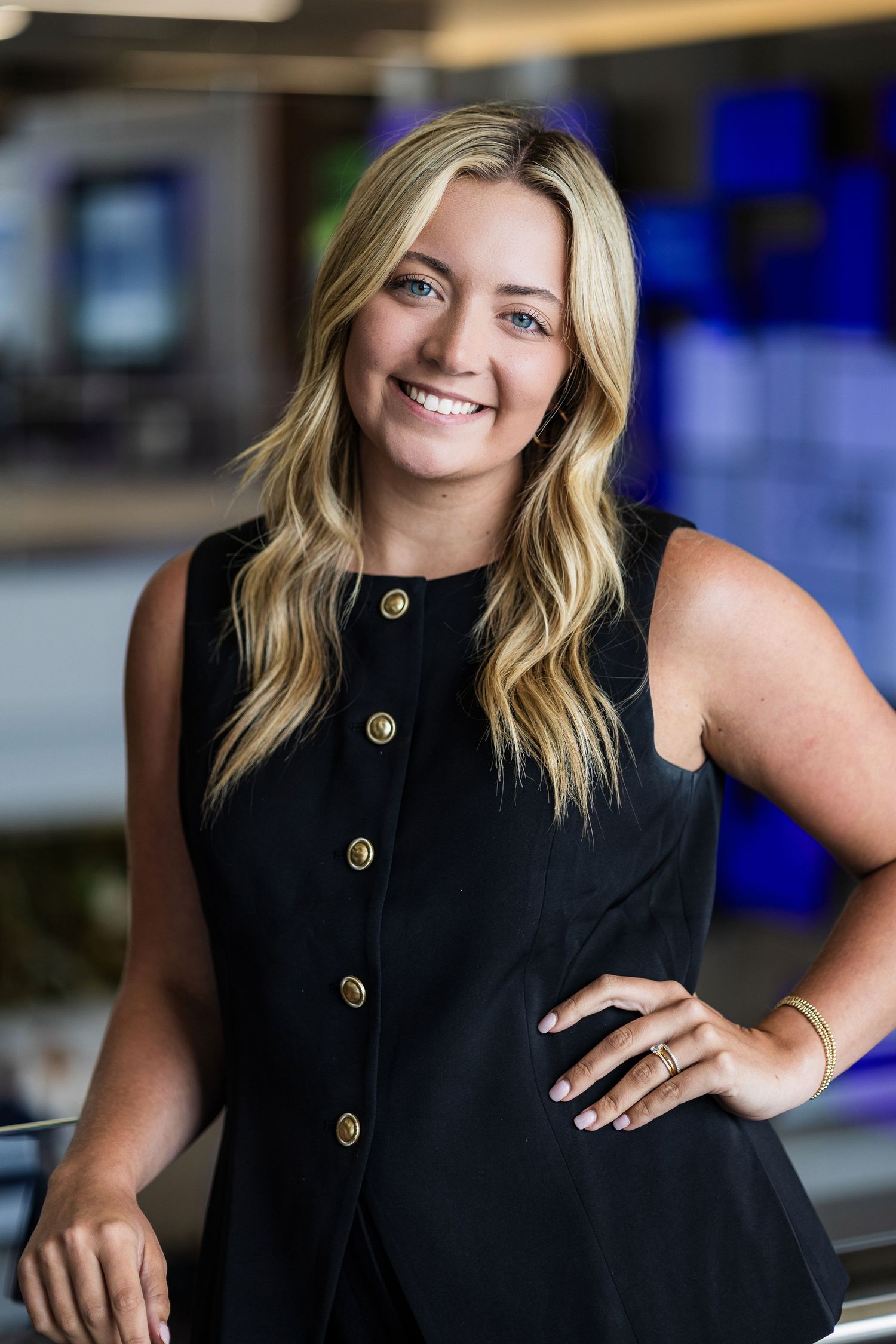 Professional headshot of a person in a sleek black sleeveless button-up top posing confidently in an office setting.