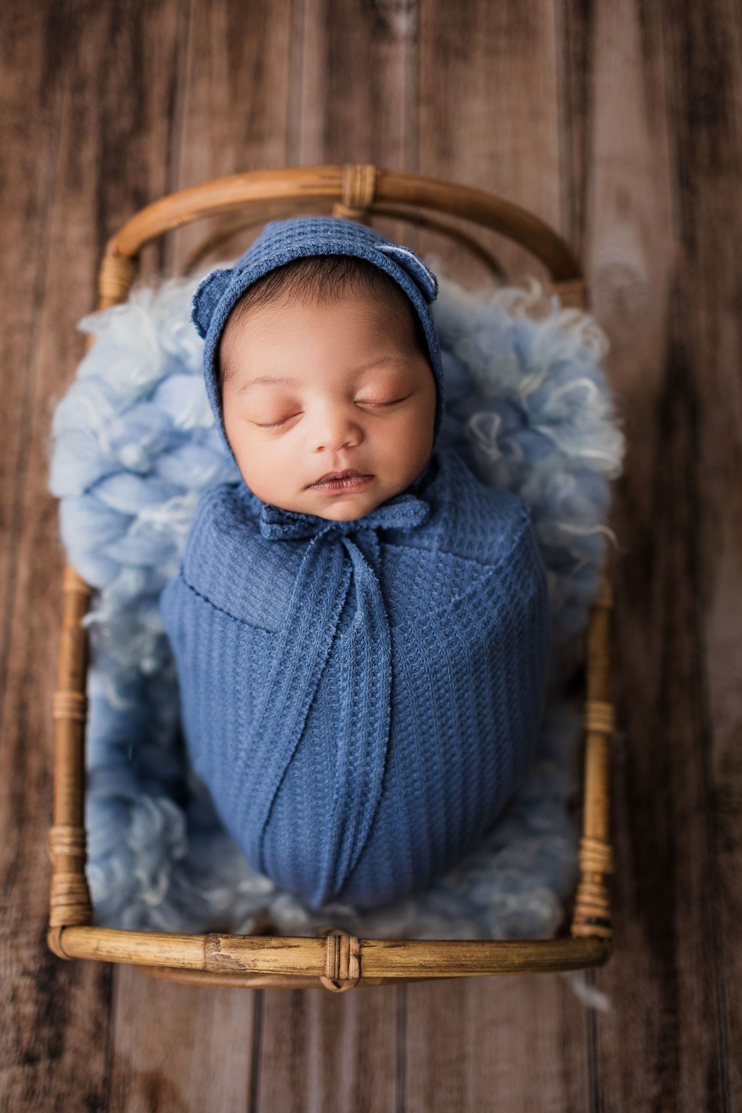 Sleeping newborn wrapped in soft blue knit blanket rests peacefully in wooden basket with matching bonnet.