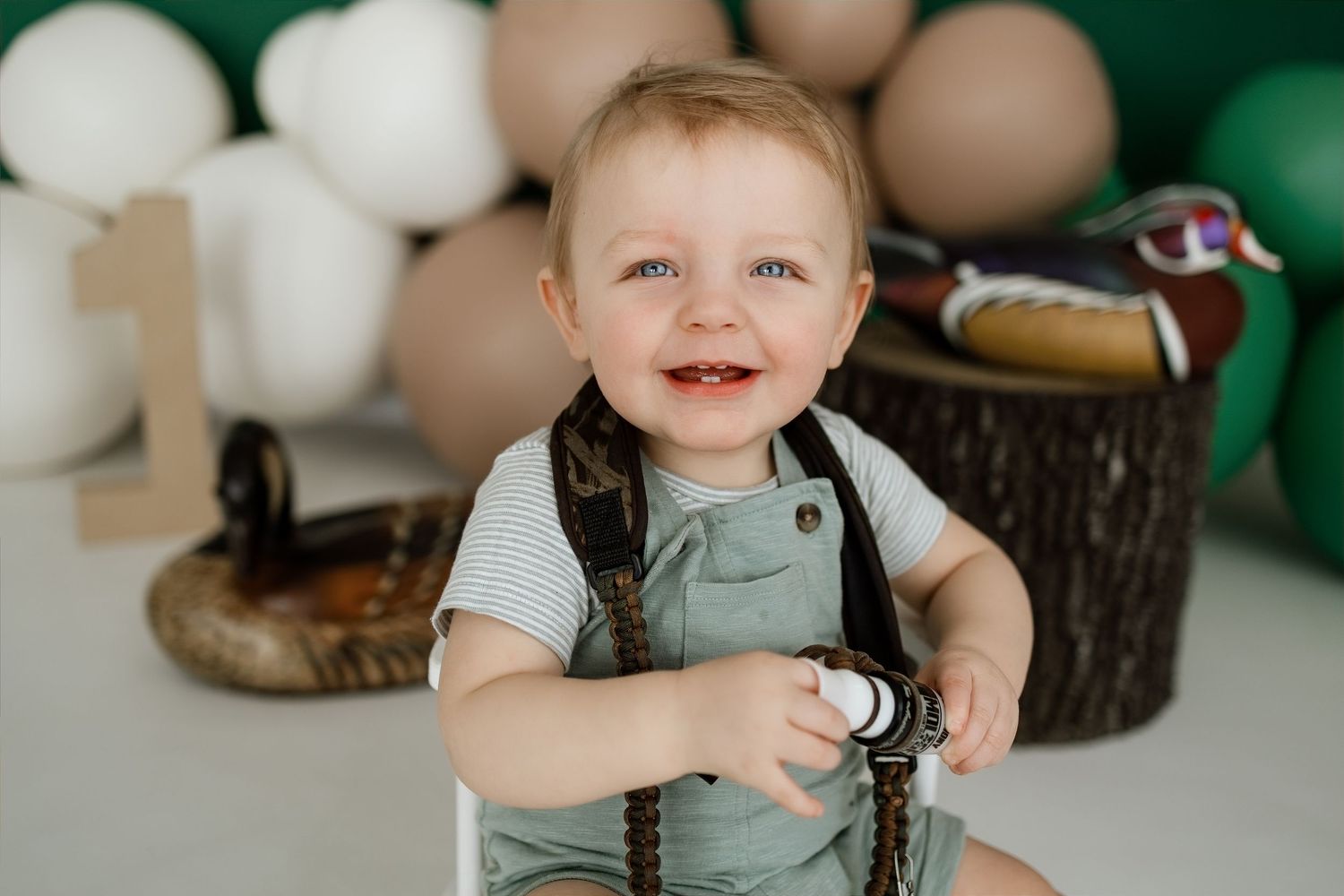 Smiling baby boy in green overalls sits by duck decor and balloons at a first birthday photo shoot.