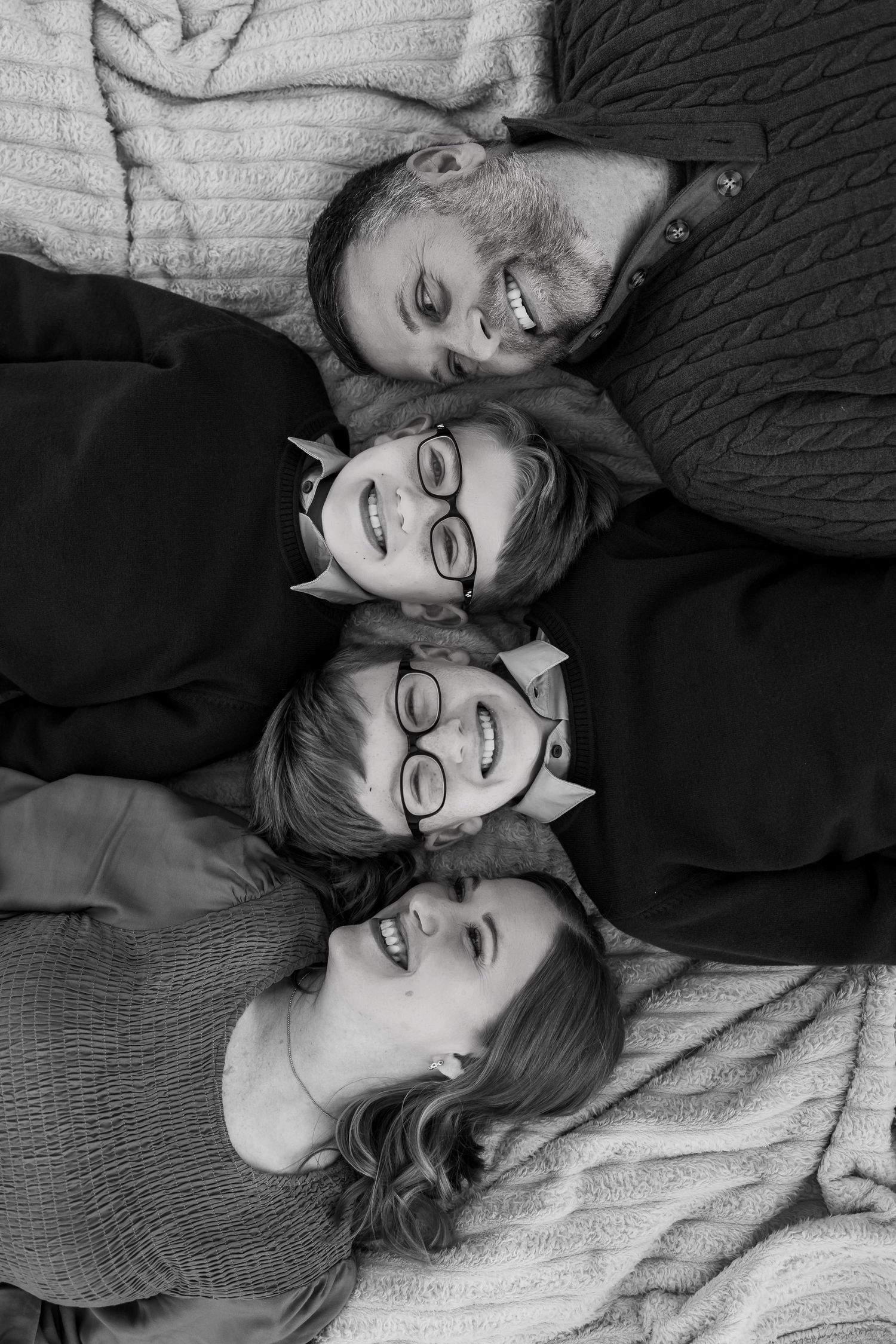 Black and white photo series of a couple lying down together and smiling while wearing glasses and cozy sweaters.