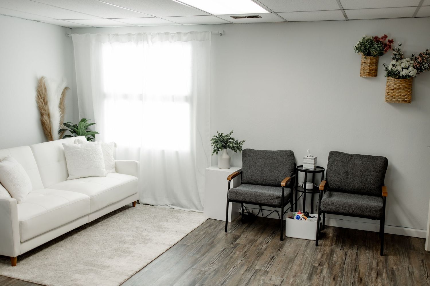 A bright, modern living room features white walls, grey seating, and natural light streaming through large windows.