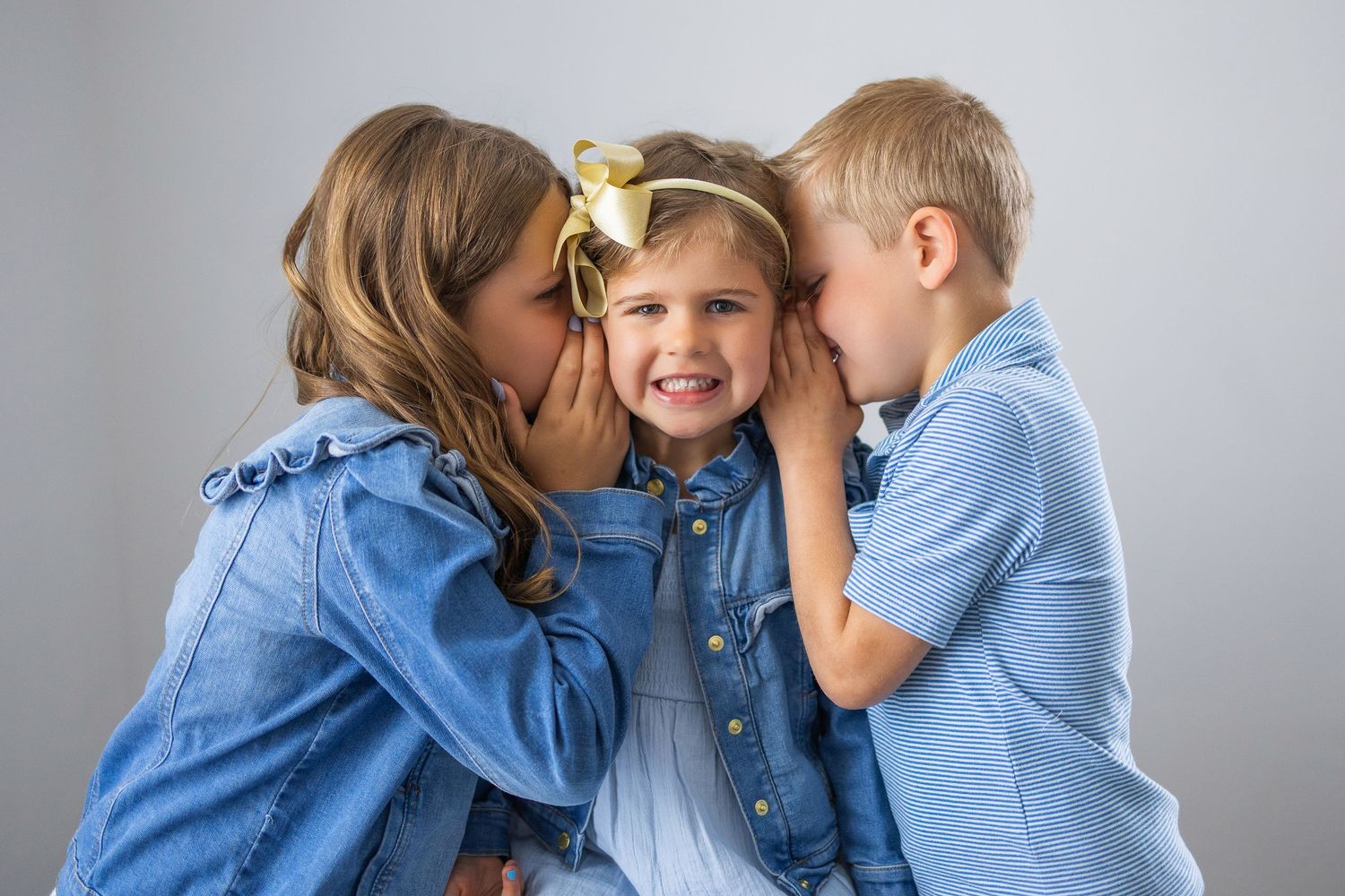 Two children in denim outfits whispering into another child's ears as they share a sweet secret moment together.