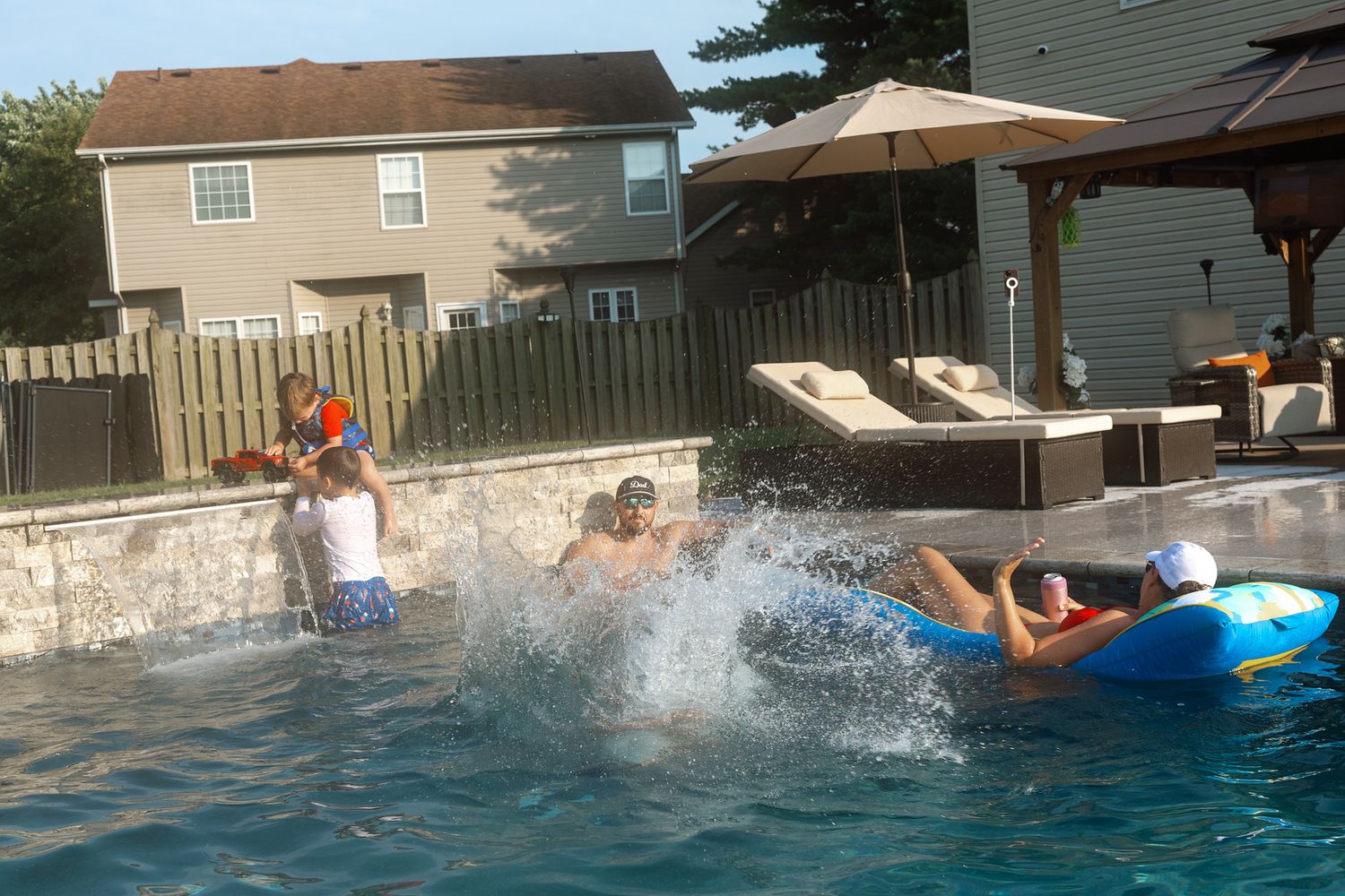Children playing in a backyard swimming pool on a sunny day, with one child on a blue float and another splashing.
