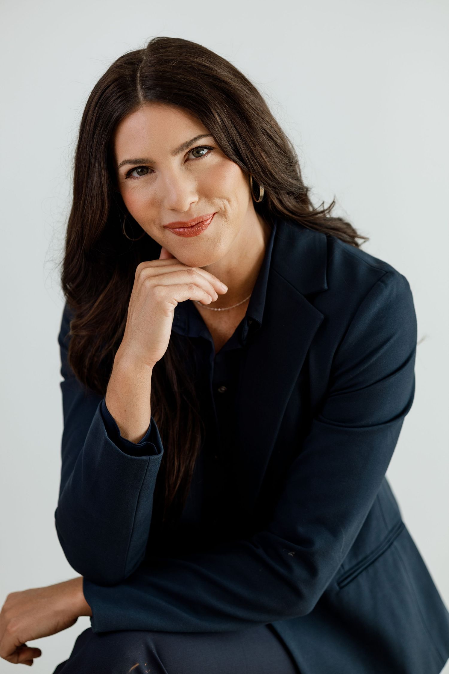 Professional woman in dark blazer smiling confidently with hand on chin against light background.