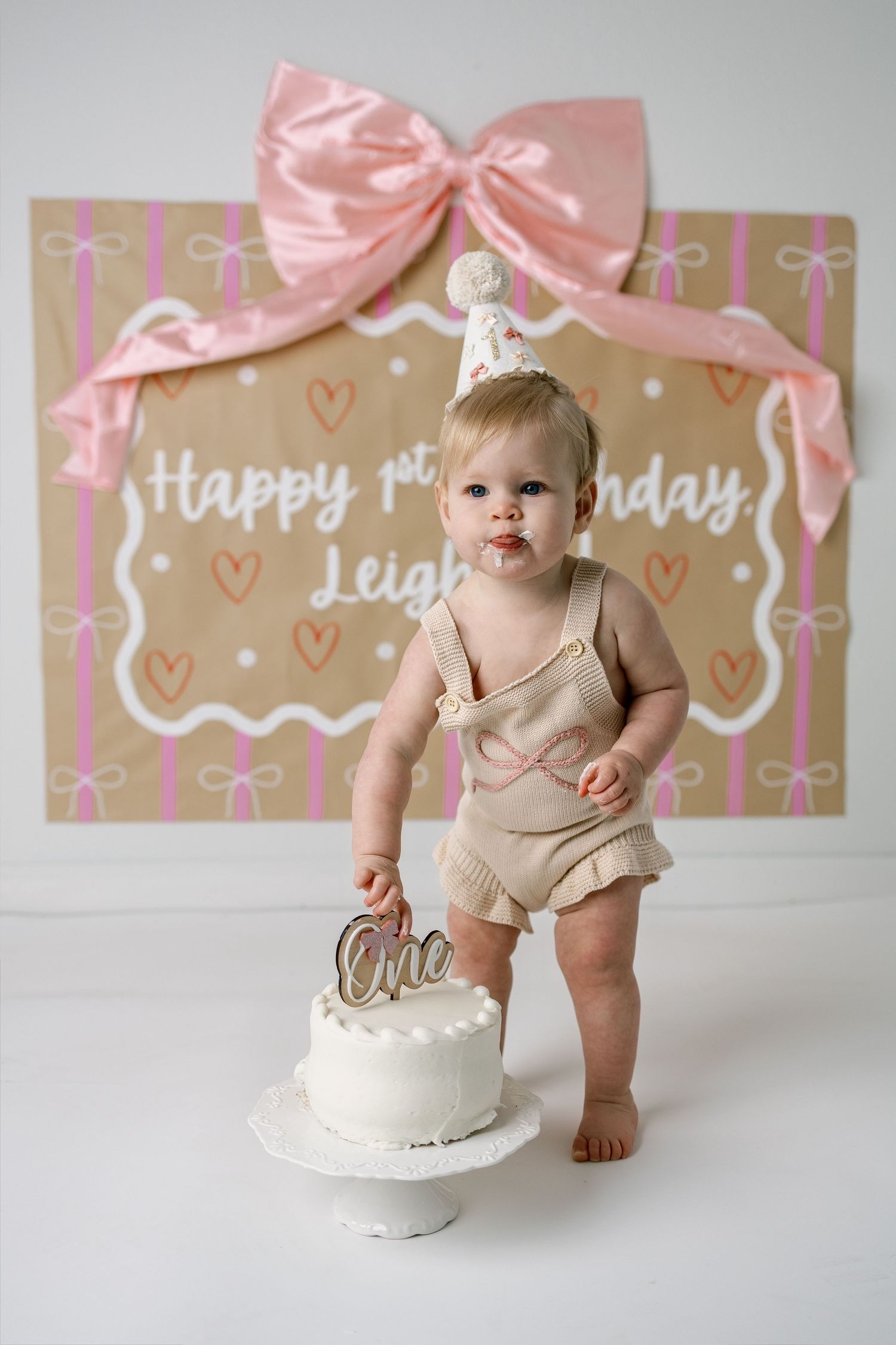 Baby in beige outfit reaching for white birthday cake with pink decorations and backdrop.