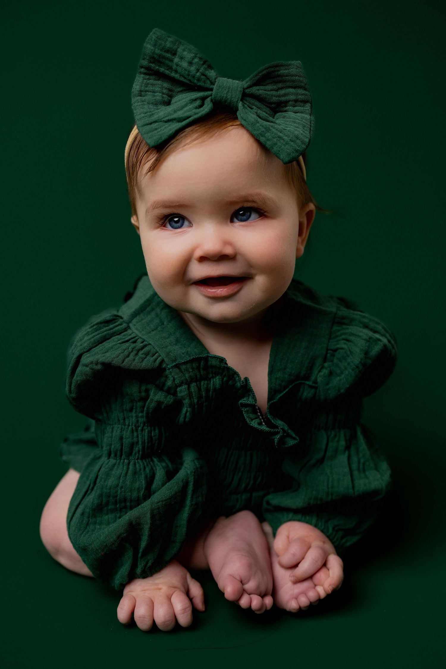 Sweet baby in emerald green dress and matching bow sits on dark green background showing tiny feet.