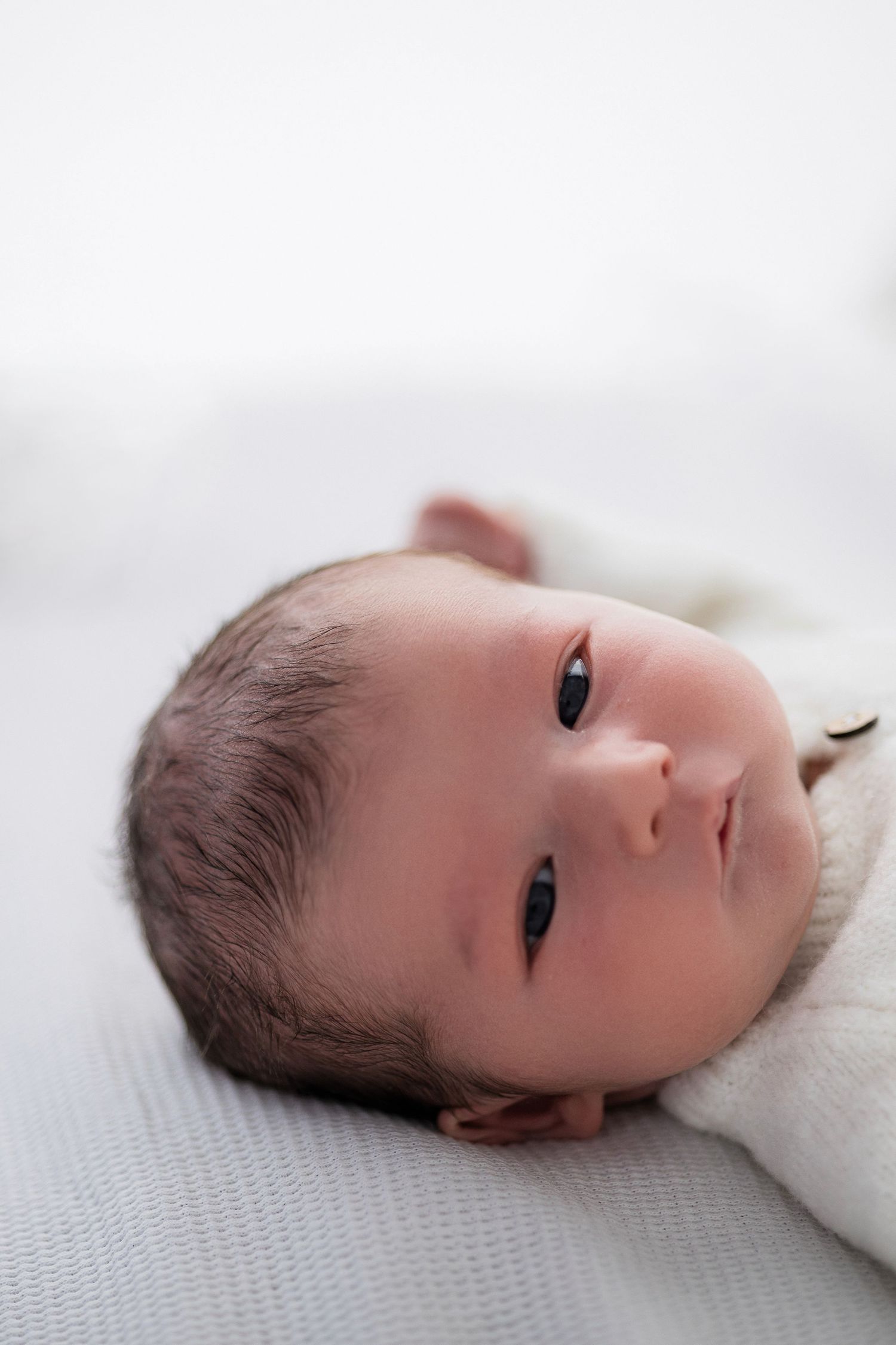 A newborn baby with soft dark hair rests on a light gray blanket while gazing upward.