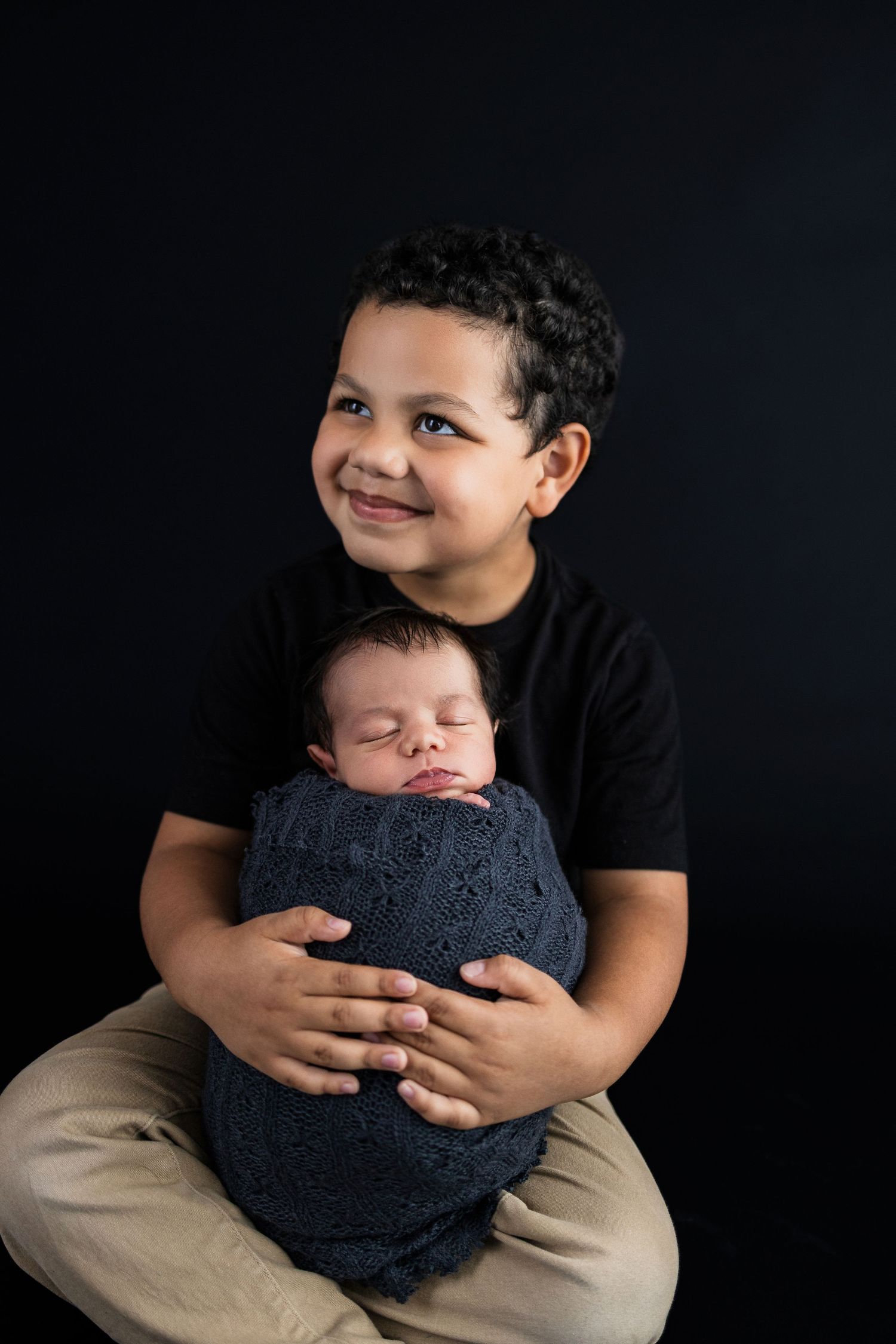 Studio portrait of a young boy in black shirt holding sleeping baby swaddled in dark blue wrap against black background.