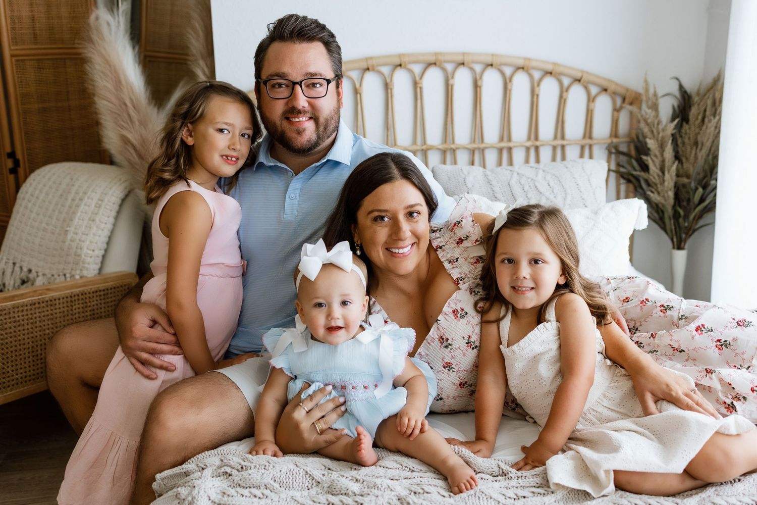 Happy family of five posing together on a bed, including parents, two young girls, and a baby in a bright bedroom.