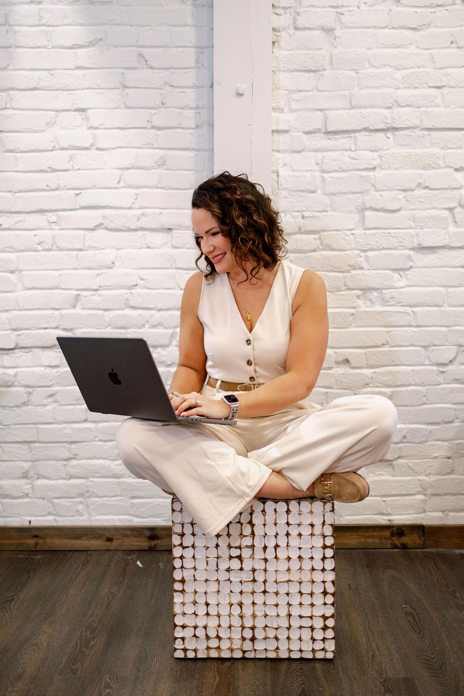 Person in white outfit working on MacBook laptop while sitting cross-legged on textured white surface against brick wall.