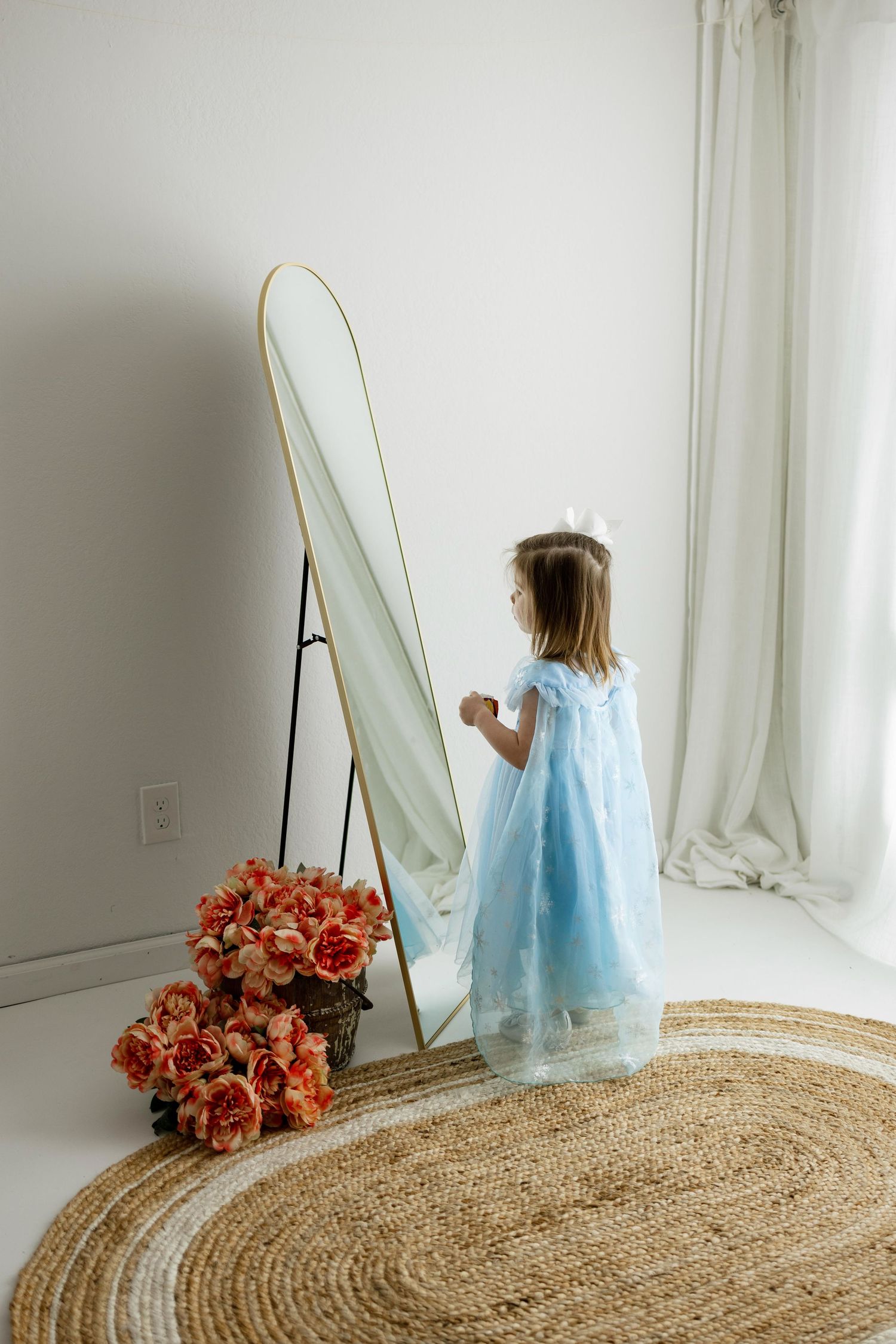 A child in a blue dress stands near a mirror with coral roses on a woven jute rug in a bright white room.