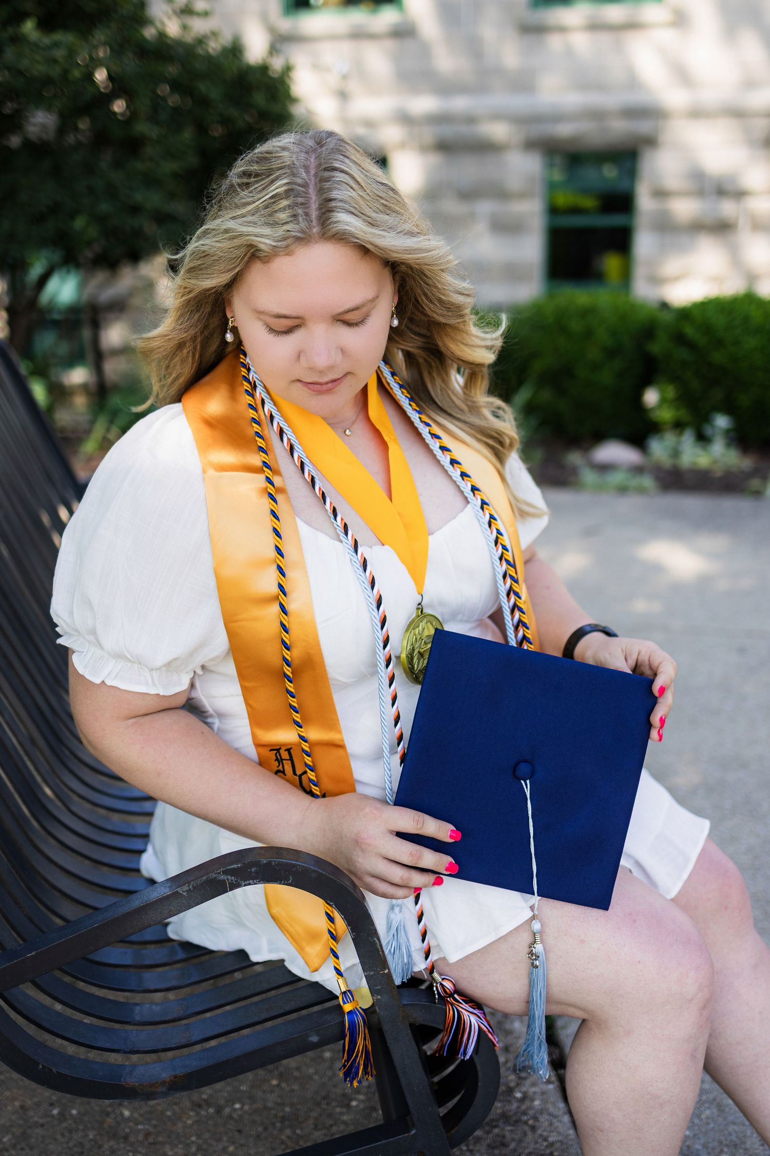 Graduate in white shirt and gold honor cords sits on bench holding blue diploma cover during outdoor ceremony.