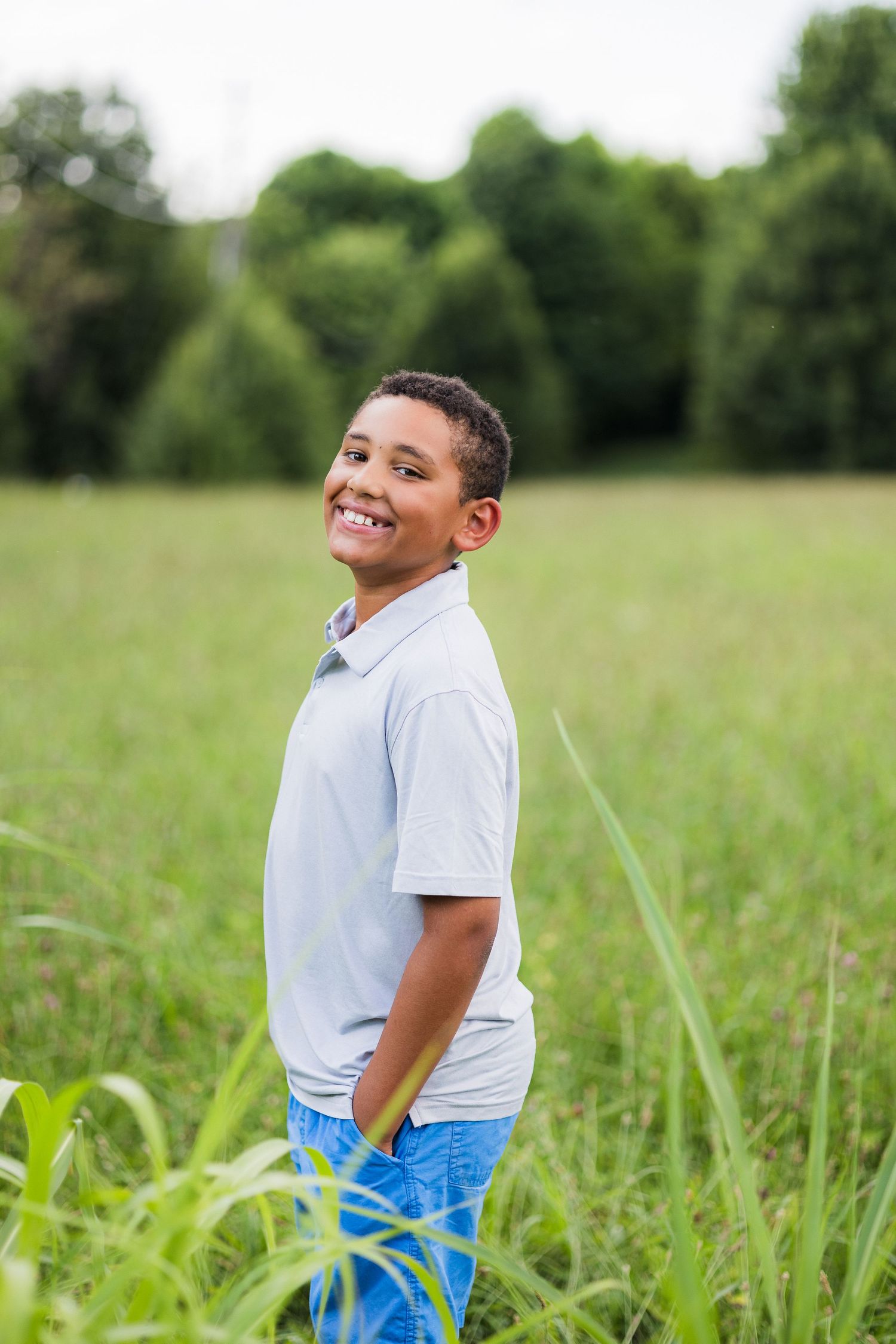 A cheerful youth wearing a light blue polo shirt and jeans stands in a tall grass field on a sunny day.