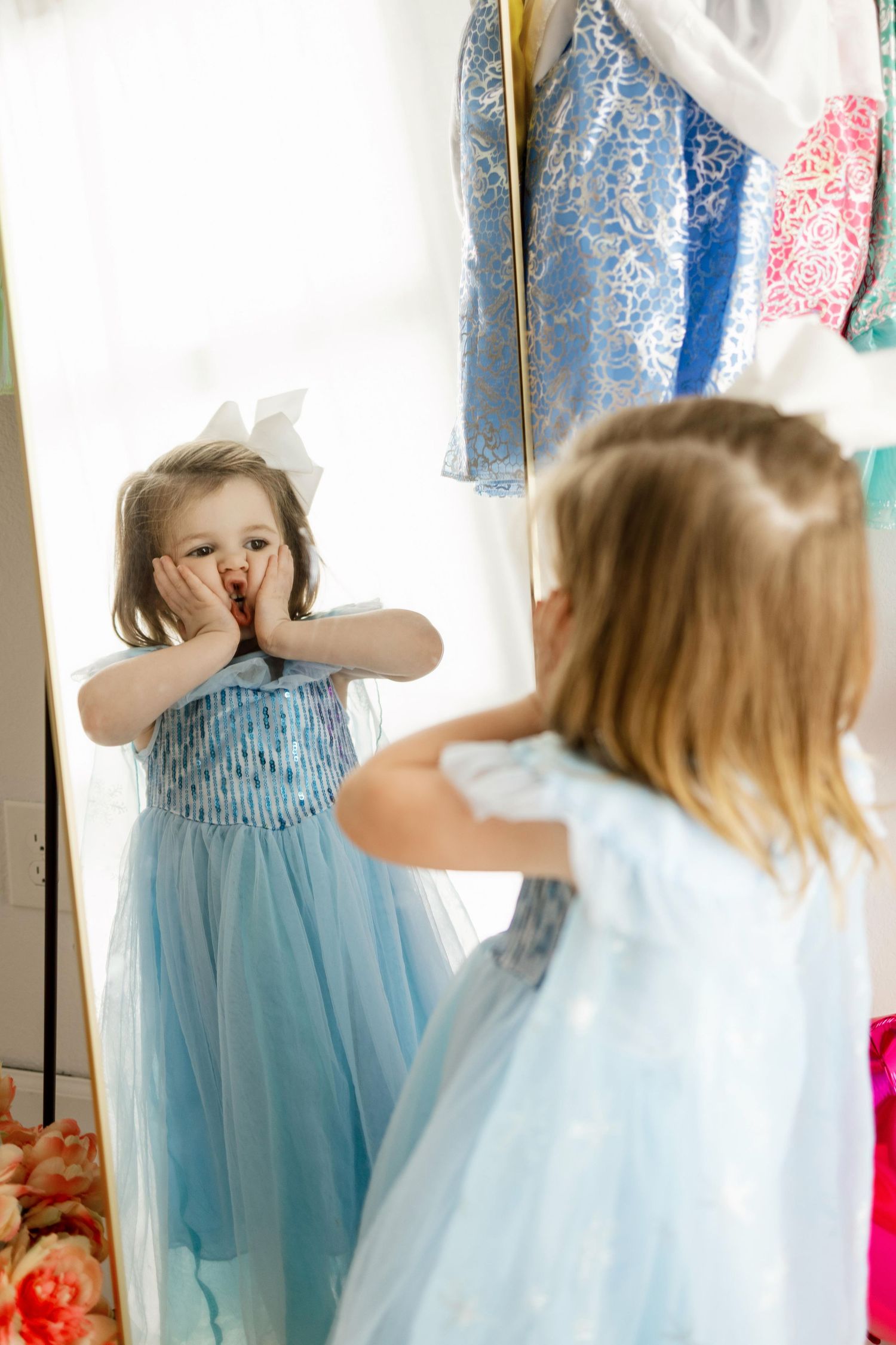 A small child in a light blue dress looks at their reflection in a mirror while playing dress-up.