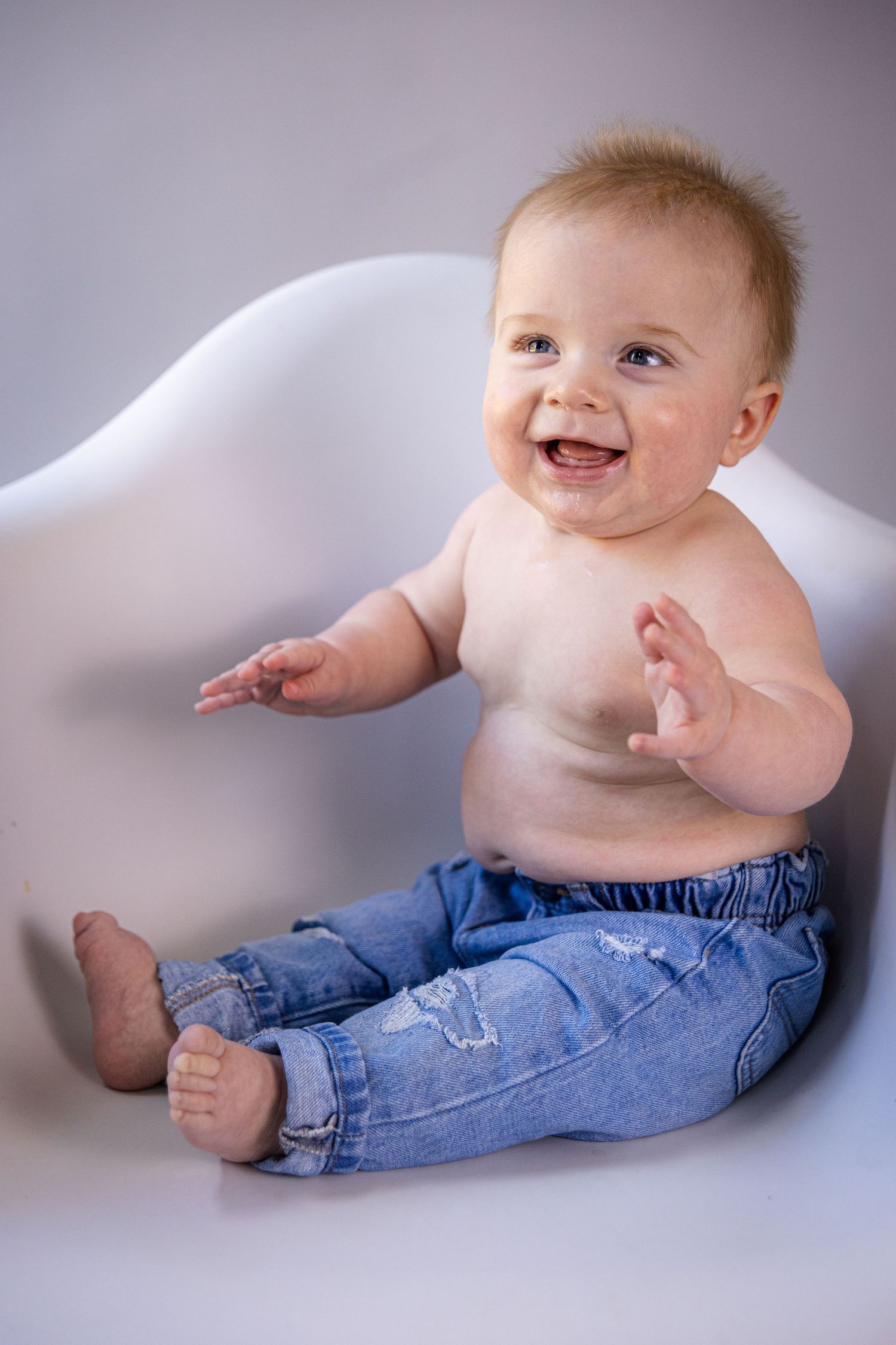 Happy baby wearing blue jeans sits on white background smiling brightly.