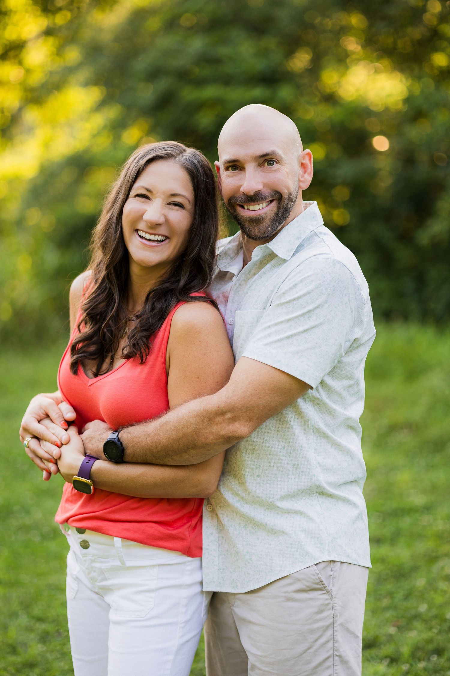 A couple embraces outdoors in casual summer attire with bright bokeh and natural lighting in the background.