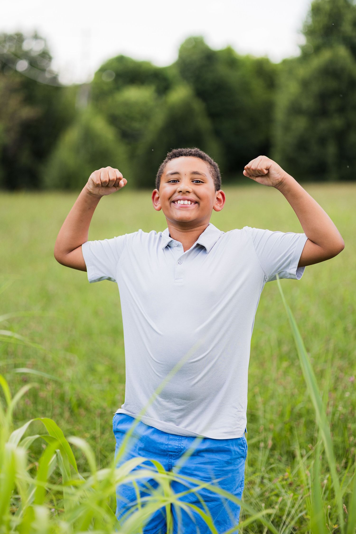 A cheerful young person playing in a grassy field while wearing a white shirt and blue shorts flexes with joy and enthusiasm.