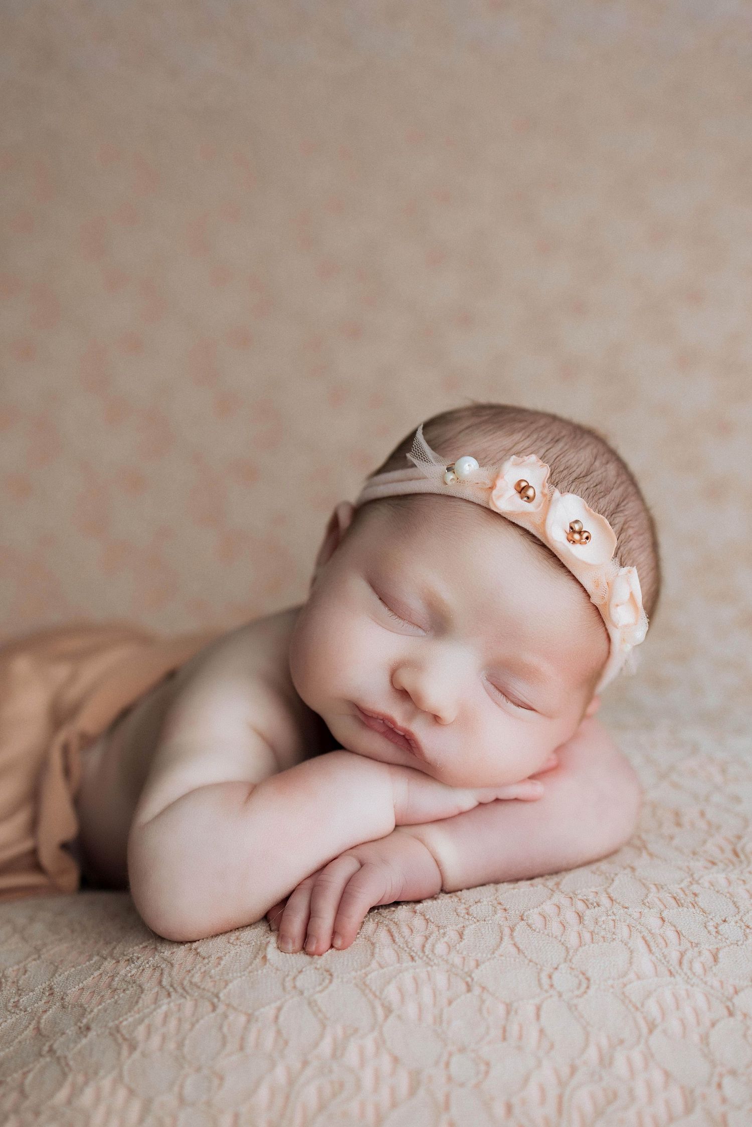 Newborn baby sleeping peacefully on textured cream-colored lace blanket wearing delicate bow headband.