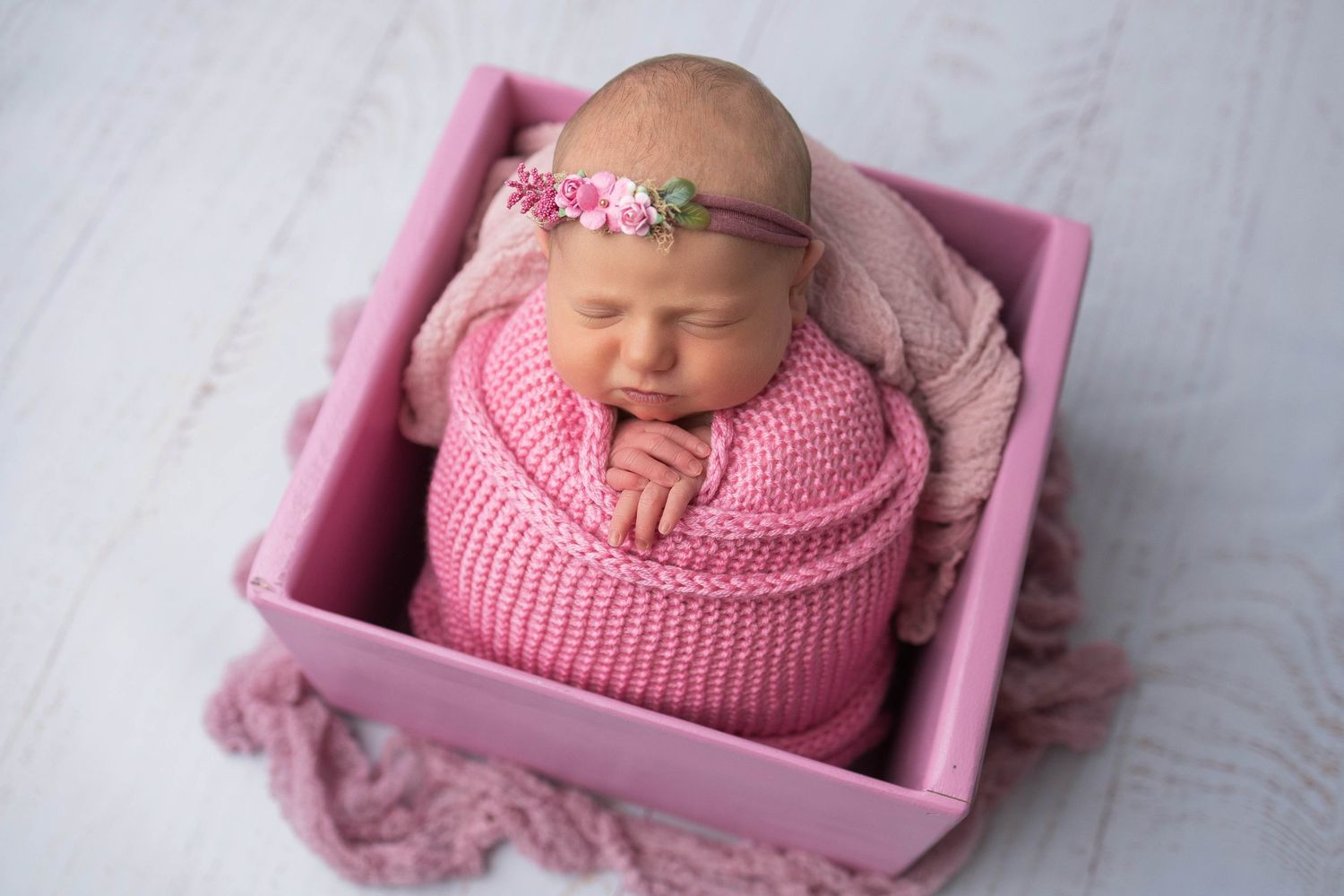 Newborn baby sleeping peacefully in pink knitted wrap inside small pink wooden box.