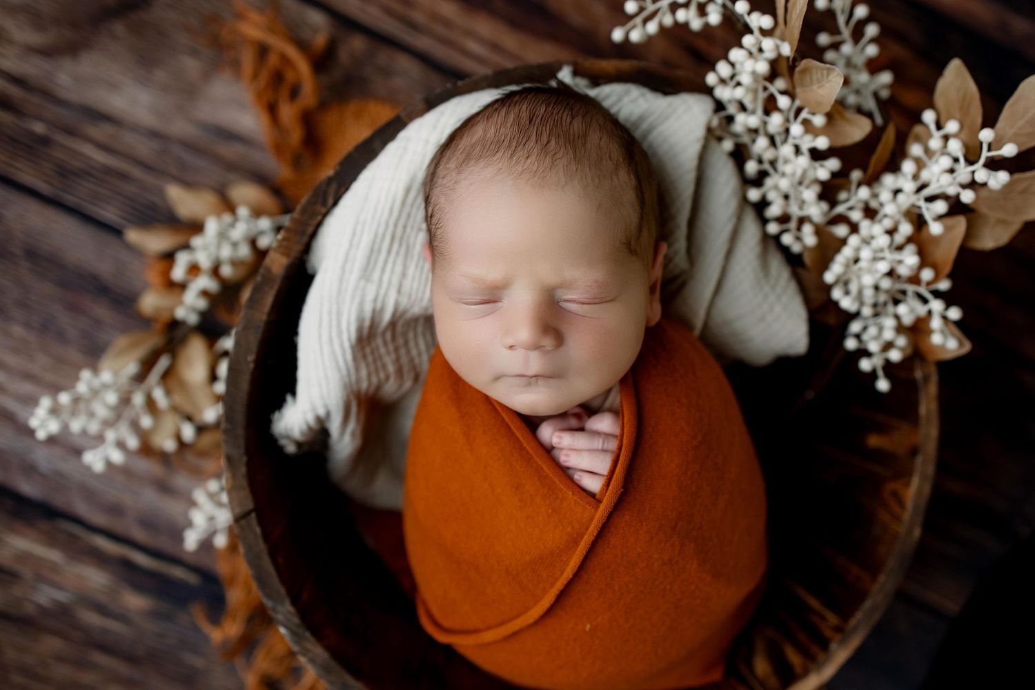 Newborn baby sleeping peacefully on brown fabric with white floral decorations against dark wooden background.