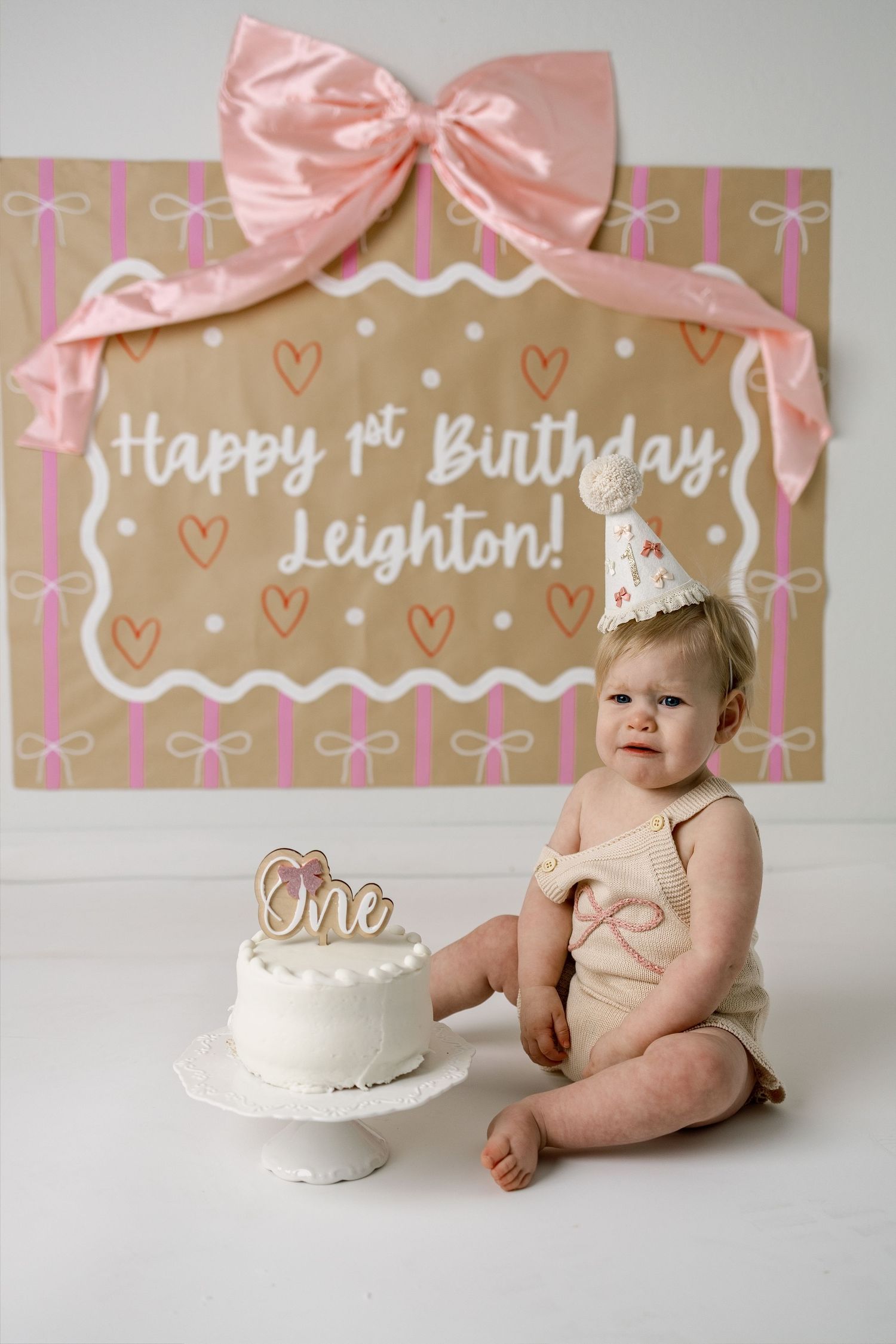First birthday setup with white cake and pink decorations against kraft paper backdrop with hearts and bows.