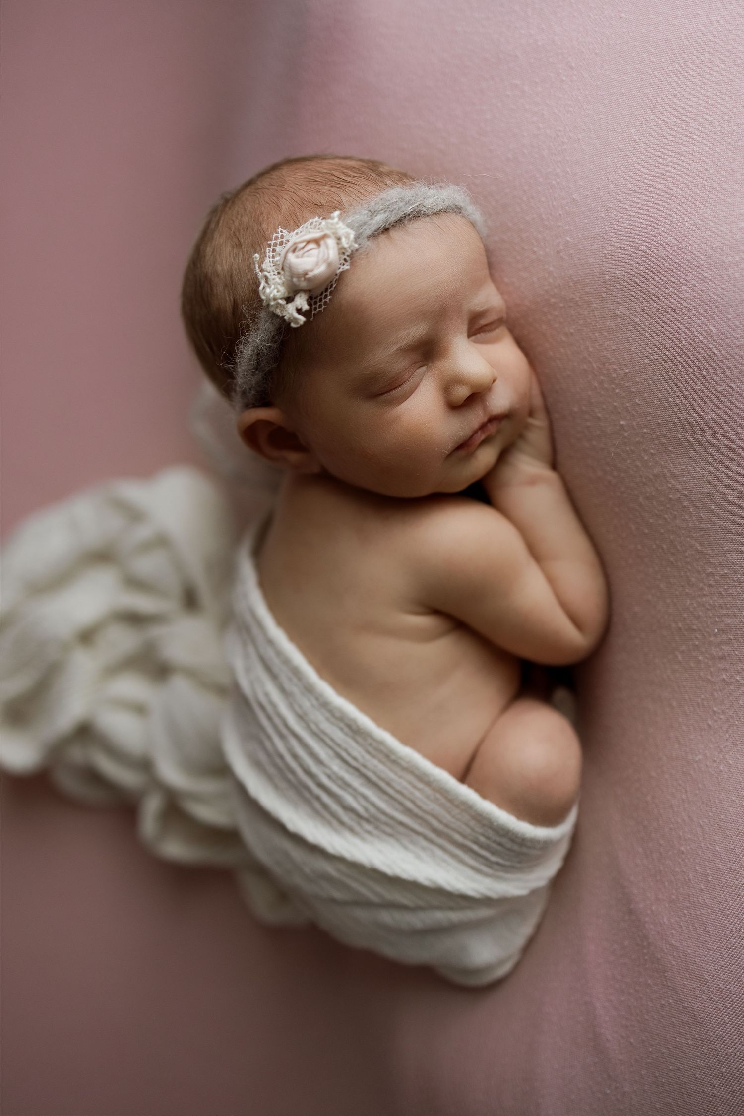 Sleeping newborn wrapped in white fabric wearing delicate lace headband against pink background.