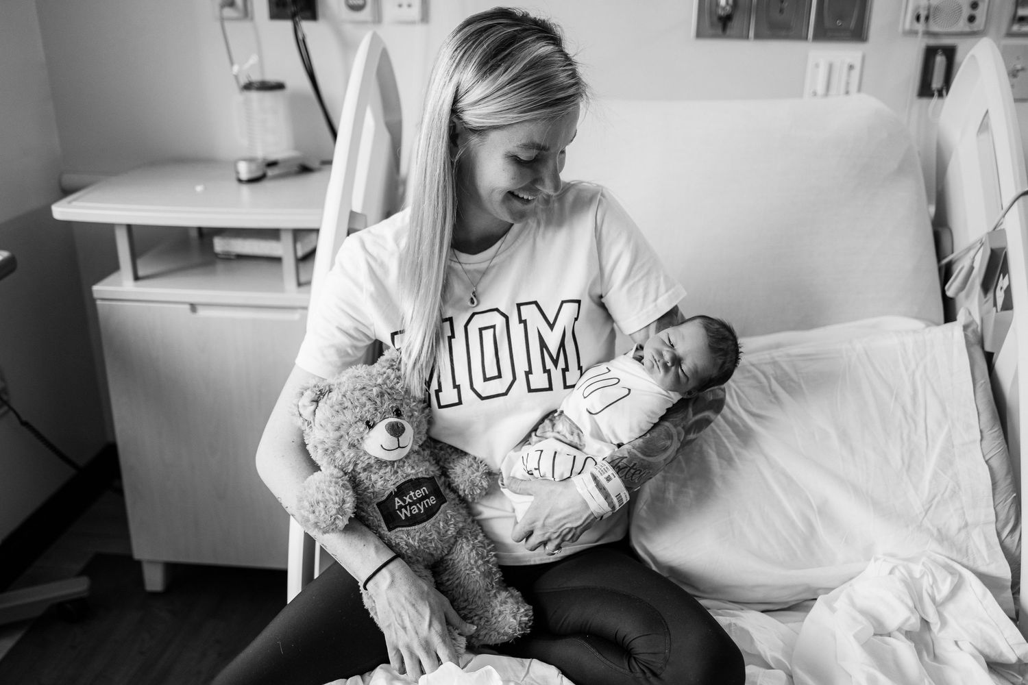 A tender black and white photo of a mother holding her newborn baby and a teddy bear in a hospital room.