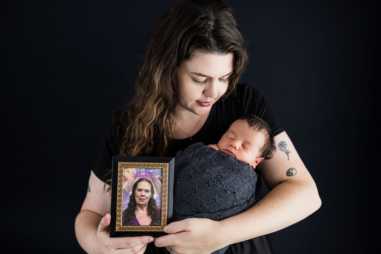 Person wearing dark clothes holds a framed photo while cradling a sleeping baby against dark background.