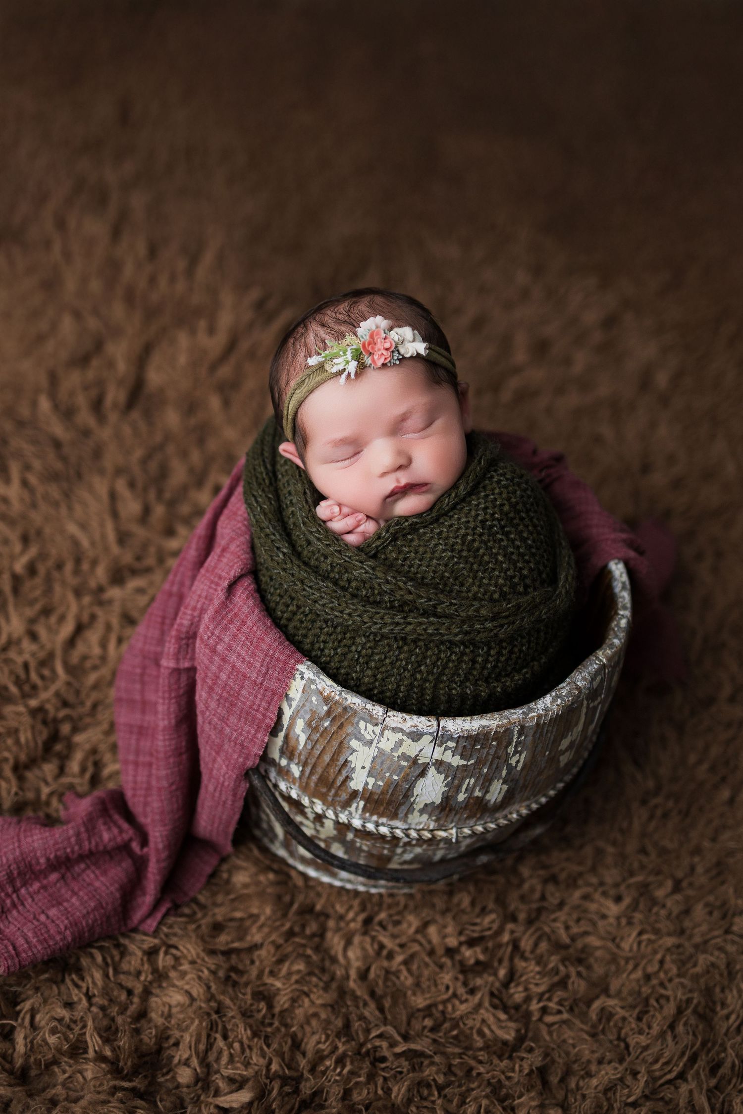 Newborn photography setup featuring a rustic basket with knitted green wrap and pink fabric against dark textured background.