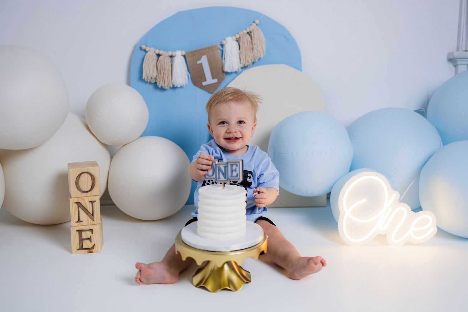 Baby sits with white birthday cake surrounded by white and blue balloons with a 'ONE' block display and banner.