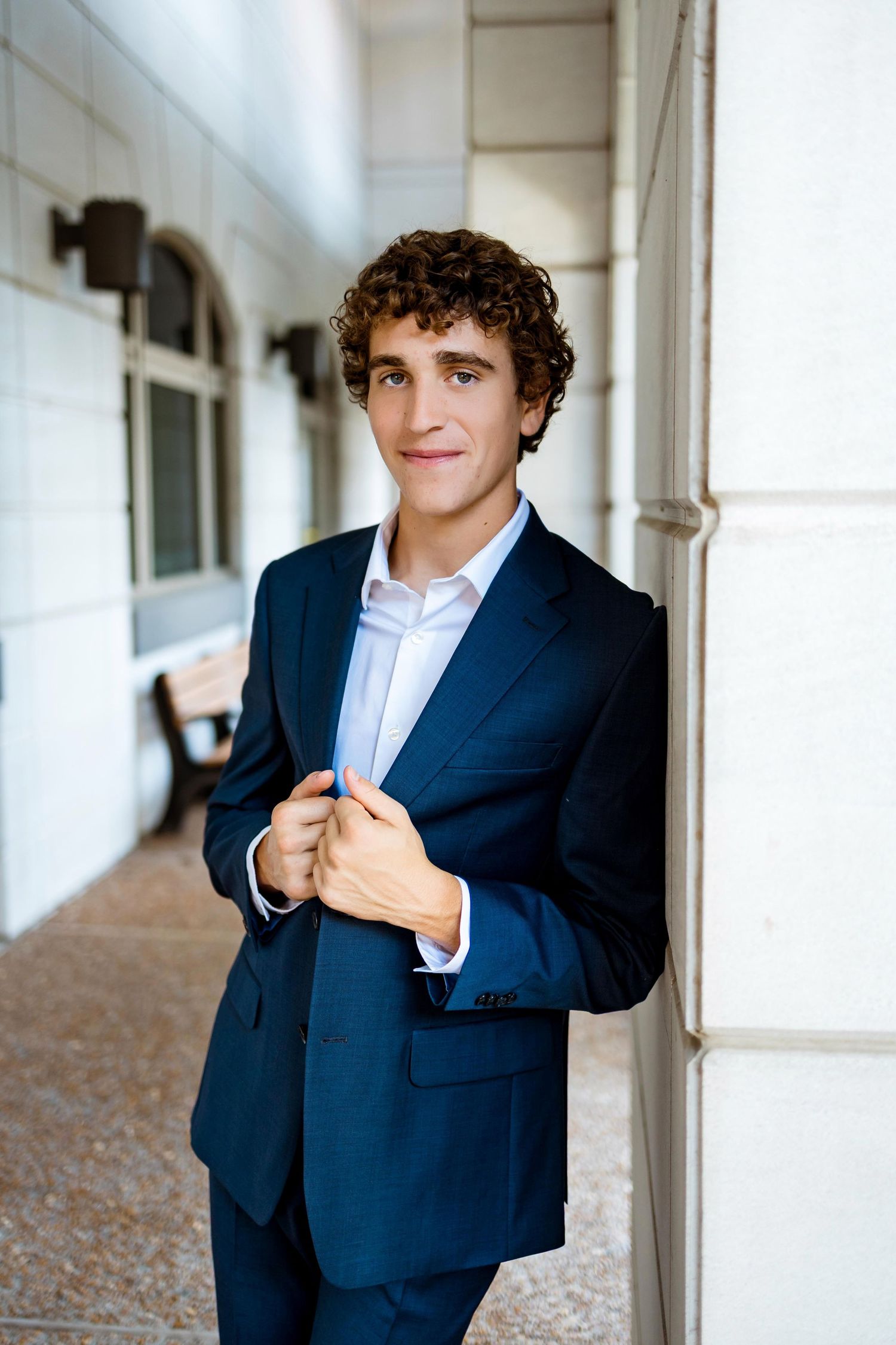 Young person in a navy blue suit and white dress shirt poses confidently against a white hallway wall.