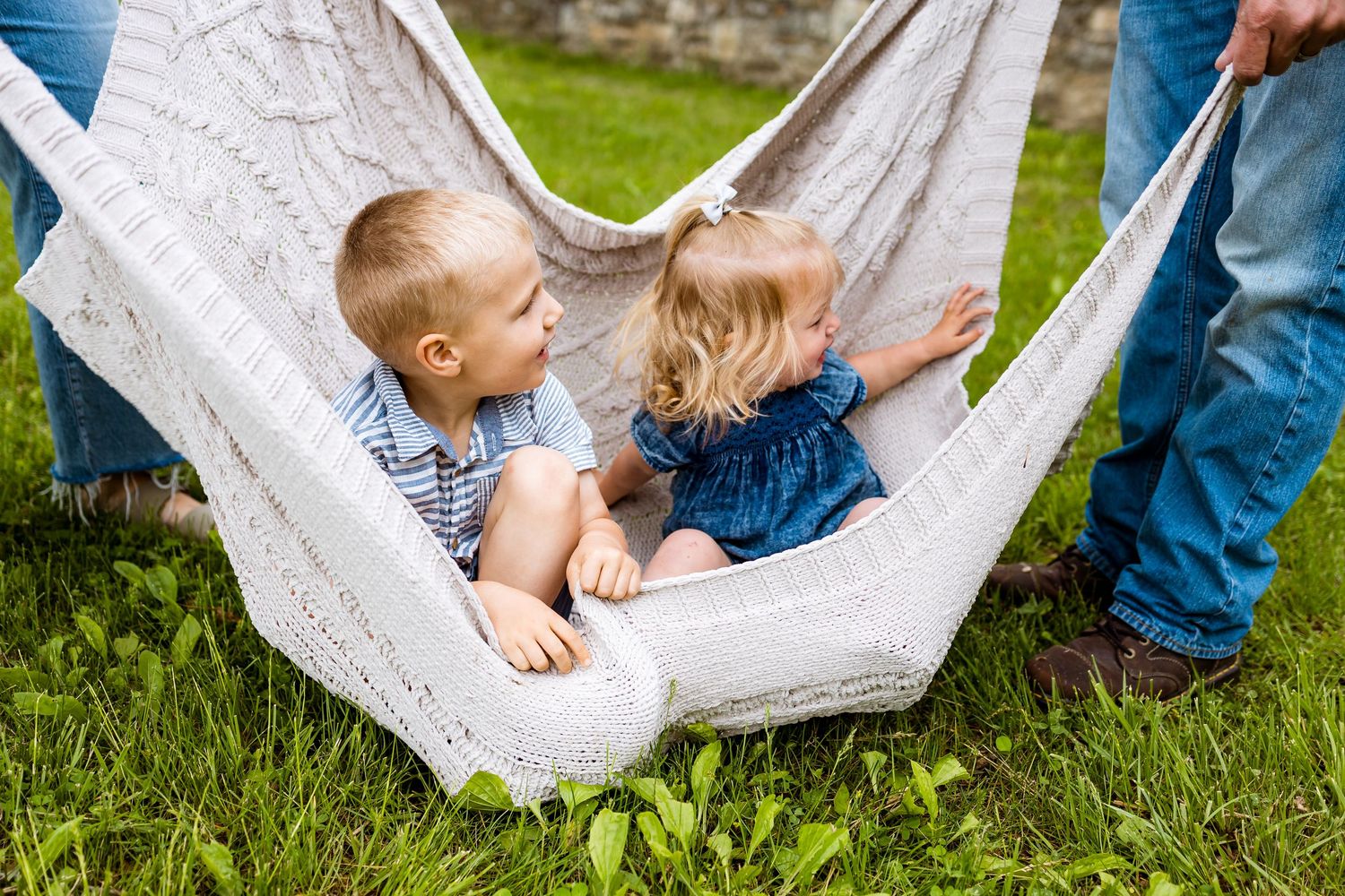 Two young children playing together in a white hammock outdoors.