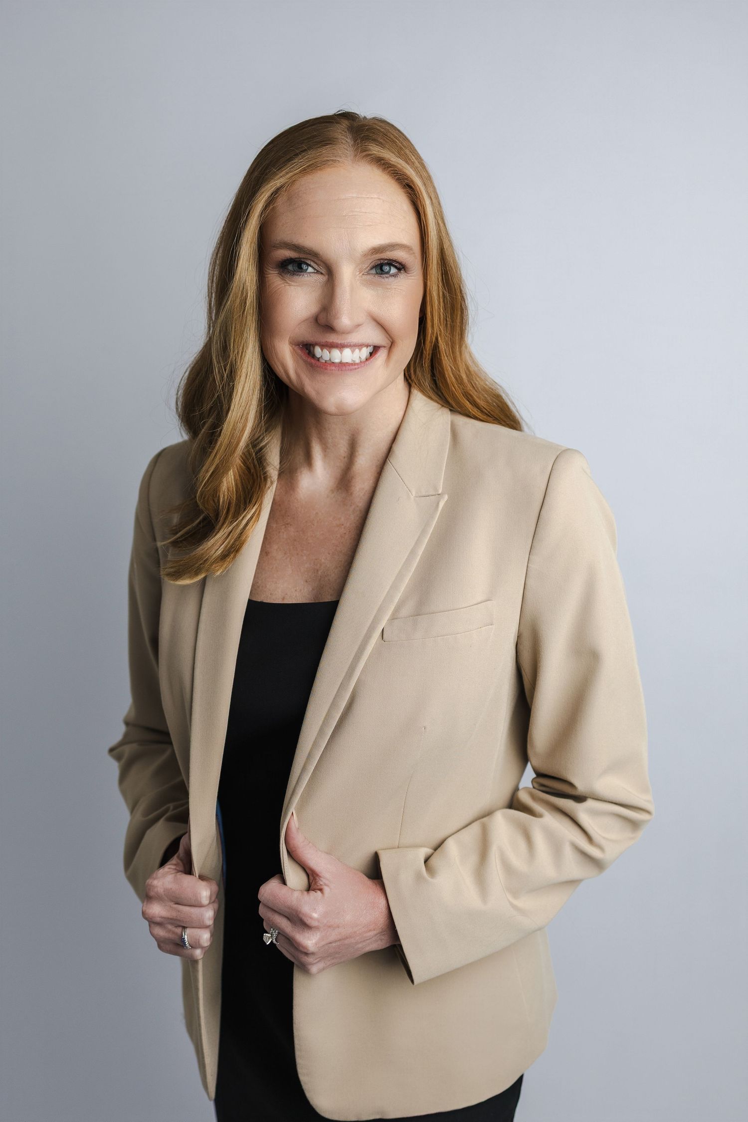 Professional headshot of person in beige blazer and black top against grey backdrop.