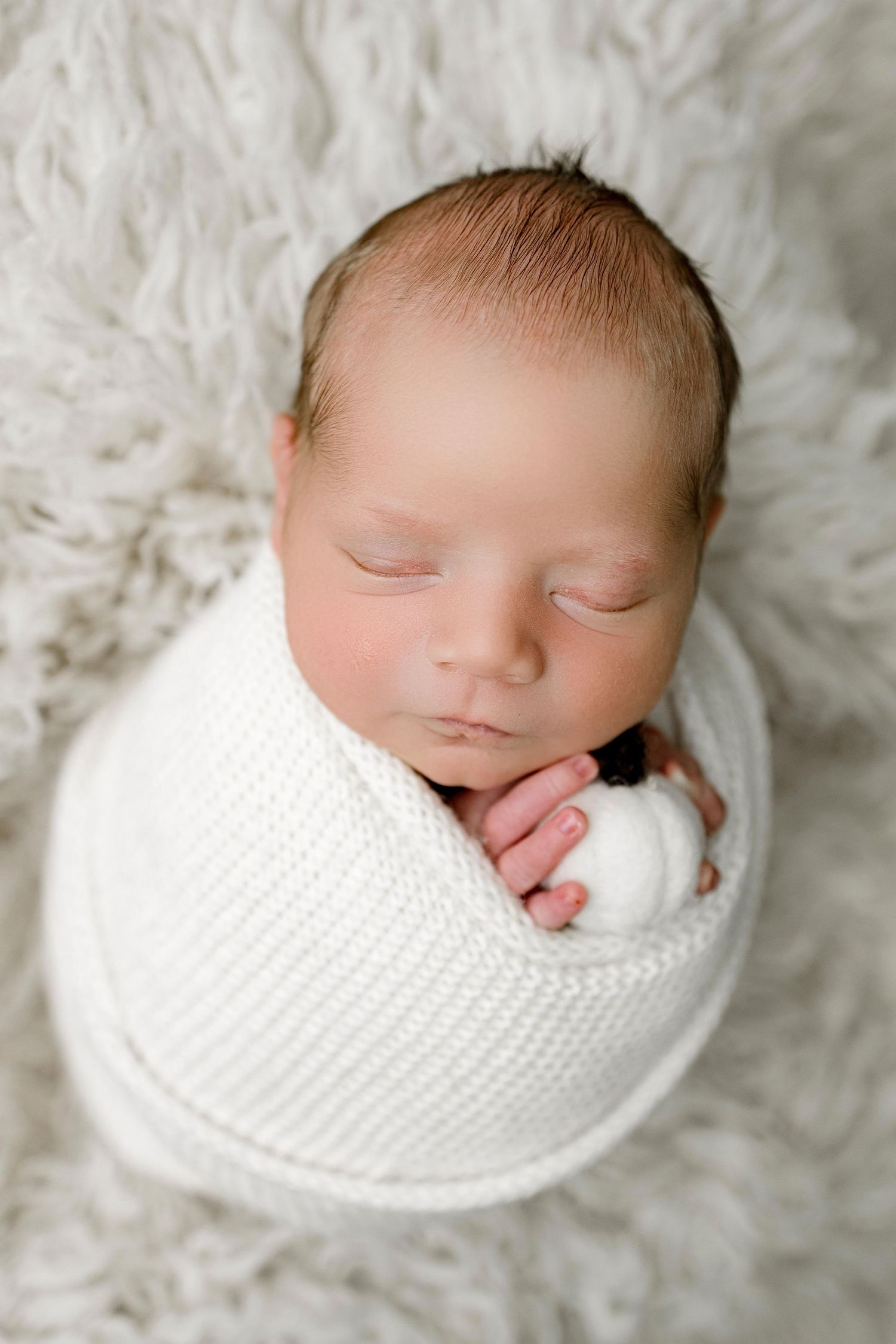Peaceful sleeping newborn baby swaddled in white knit wrap on soft furry background.