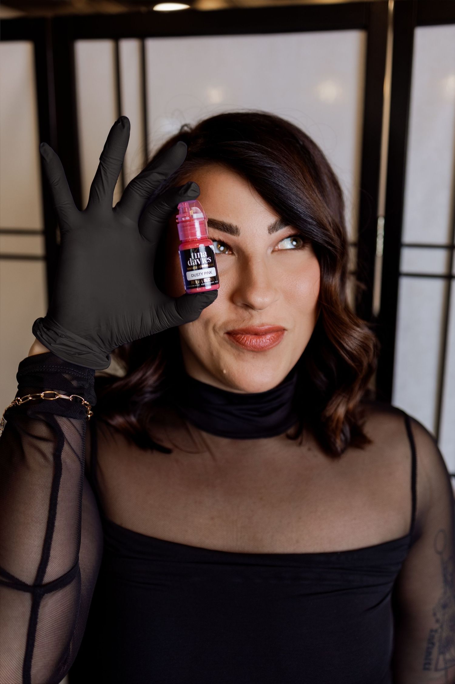 A person wearing black gloves and sheer top holds up a small pink bottle against a dark background.