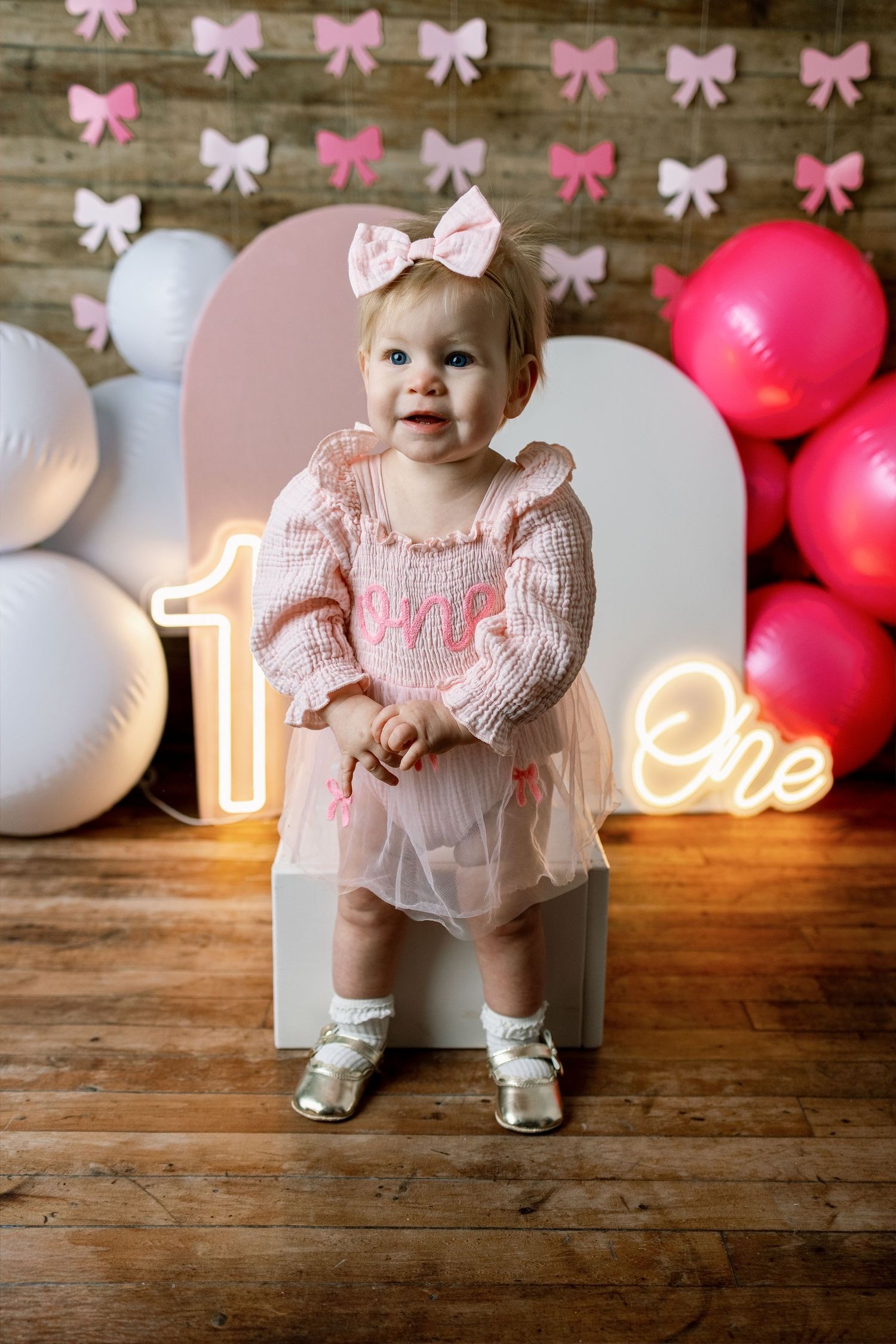 First birthday party setup with pink and white decor, balloons, bow decorations and neon number one sign on wooden floor.