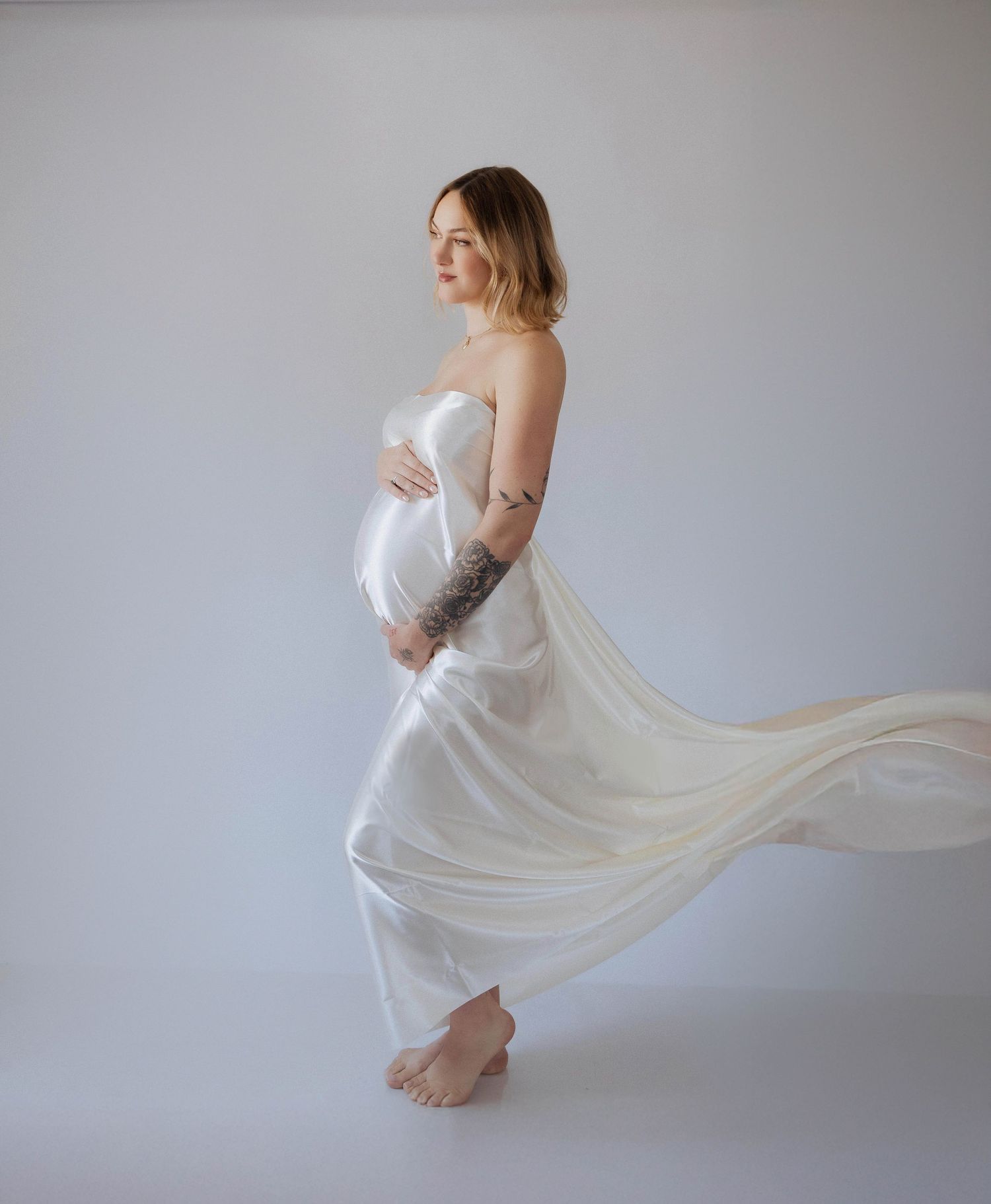Ethereal maternity photo with flowing white dress creating elegant movement against light grey background.