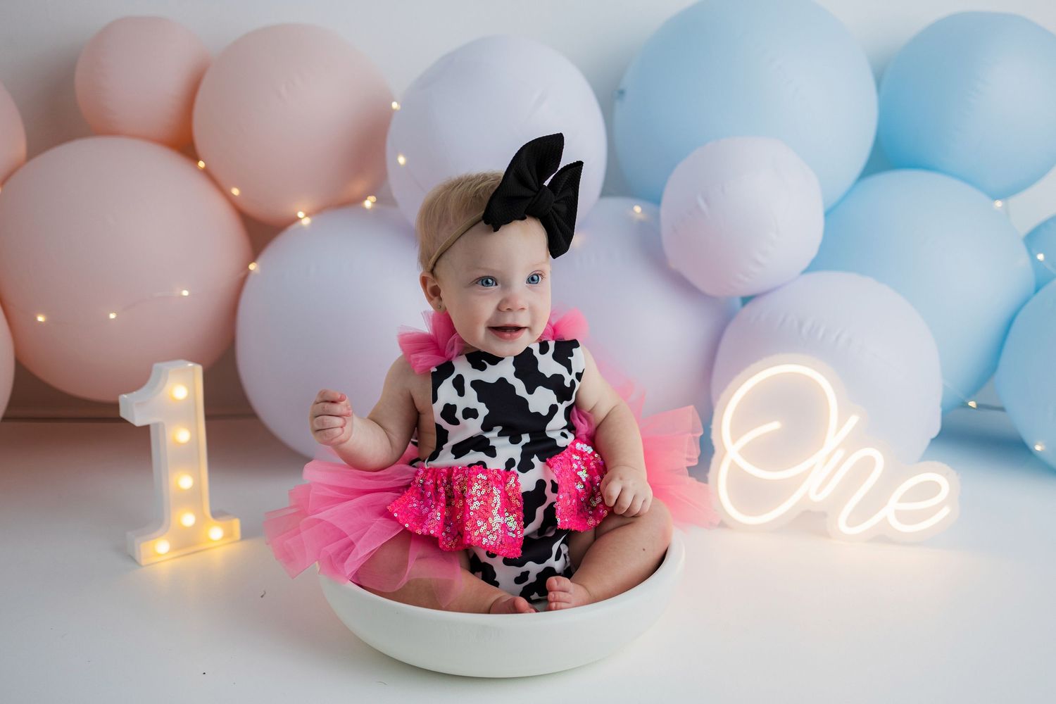 First birthday celebration scene with pink tutu outfit against pastel balloon backdrop and illuminated 'One' sign.