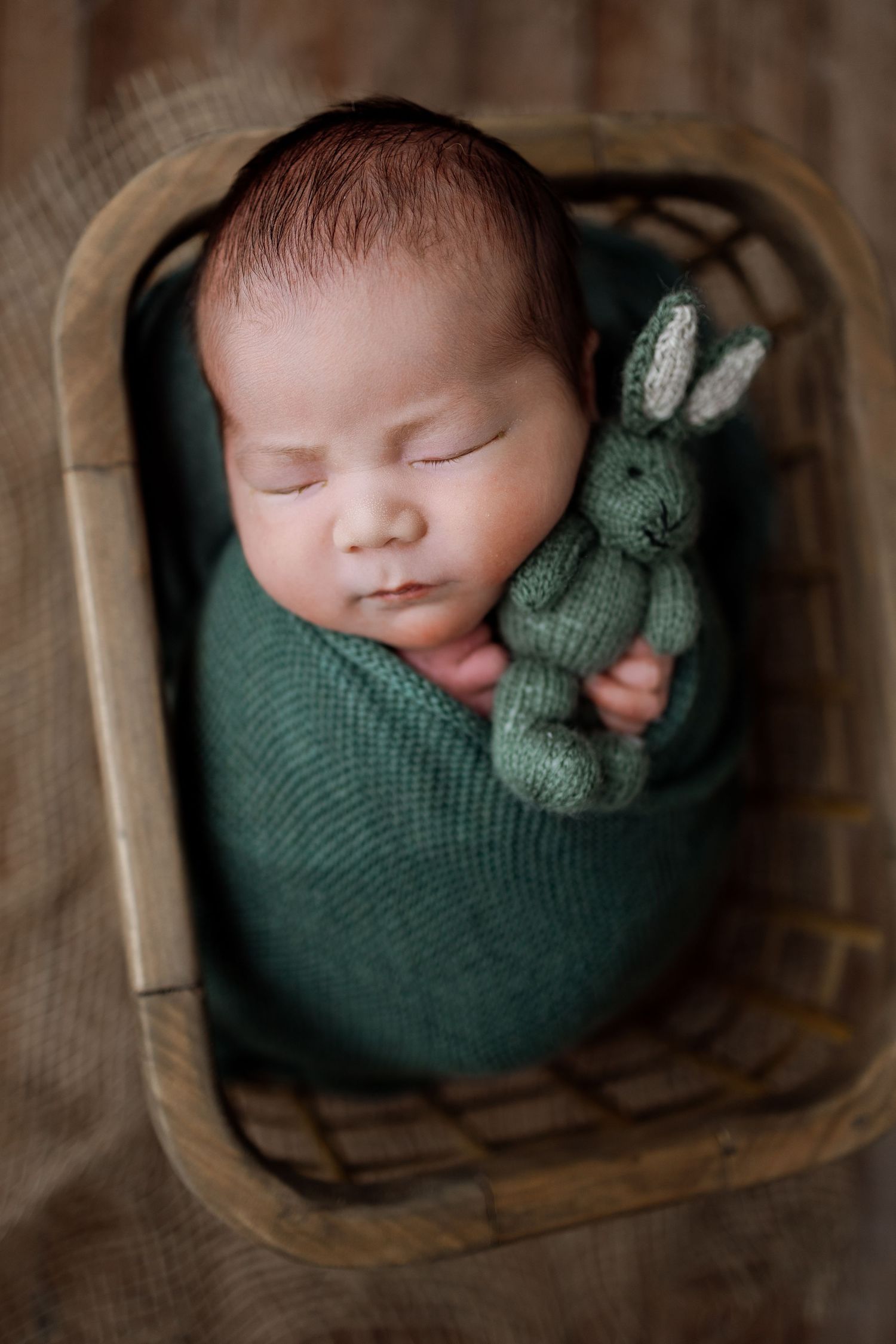 Sleeping newborn wrapped in green knit blanket rests peacefully in wooden basket.