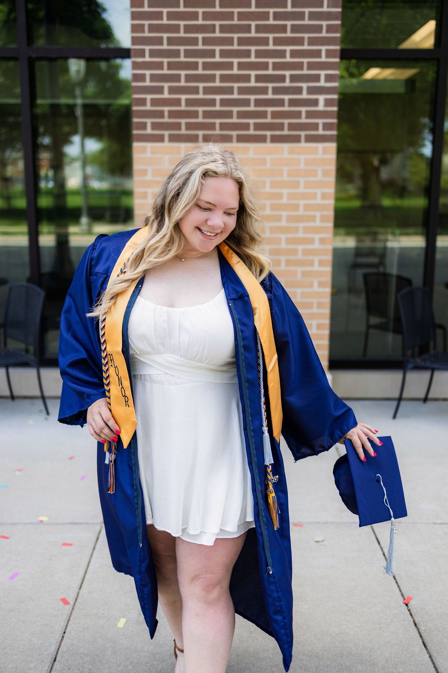 Graduate wearing white dress and blue graduation gown with gold honor cord smiles joyfully during outdoor photo session.