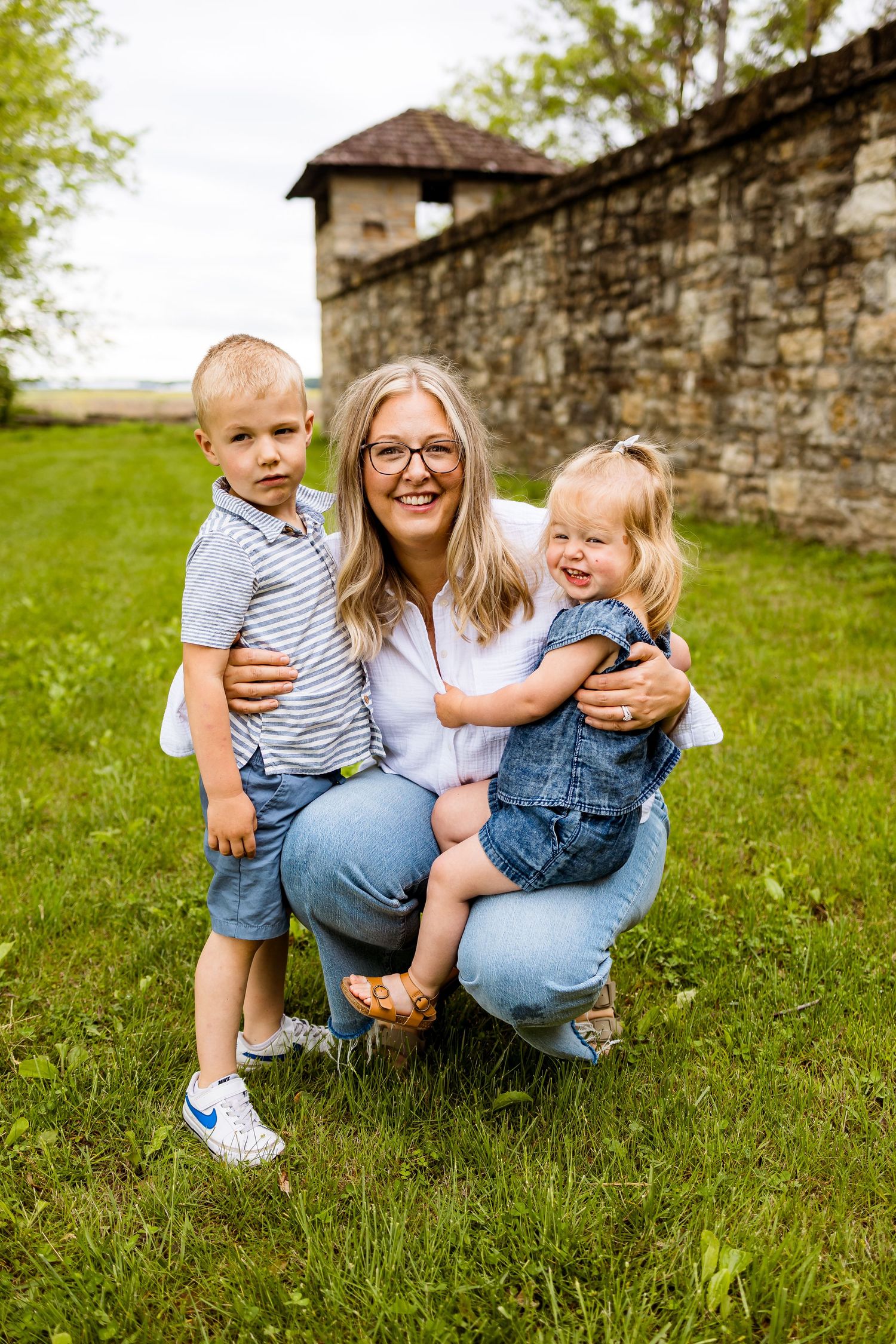 Mother kneeling on grass with two young children in casual clothing.