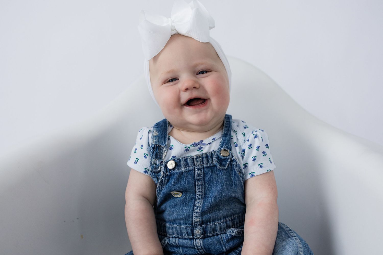 Laughing baby in denim overalls and floral shirt wearing white bow against white backdrop.