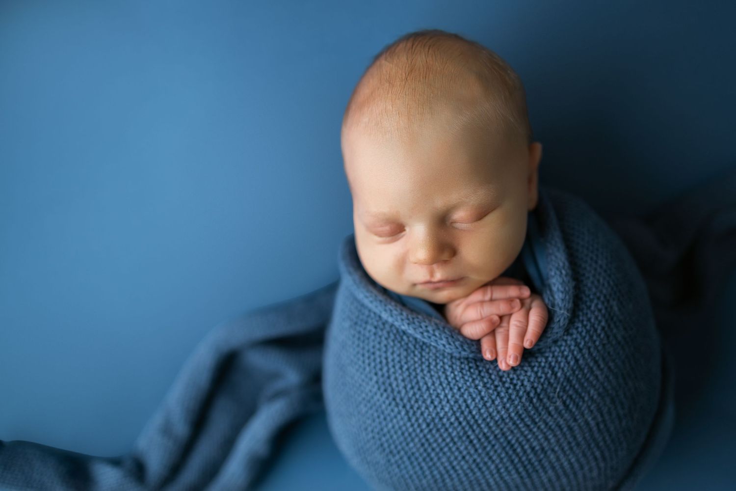 Newborn wrapped in soft blue knit swaddle against blue background in peaceful sleeping pose.